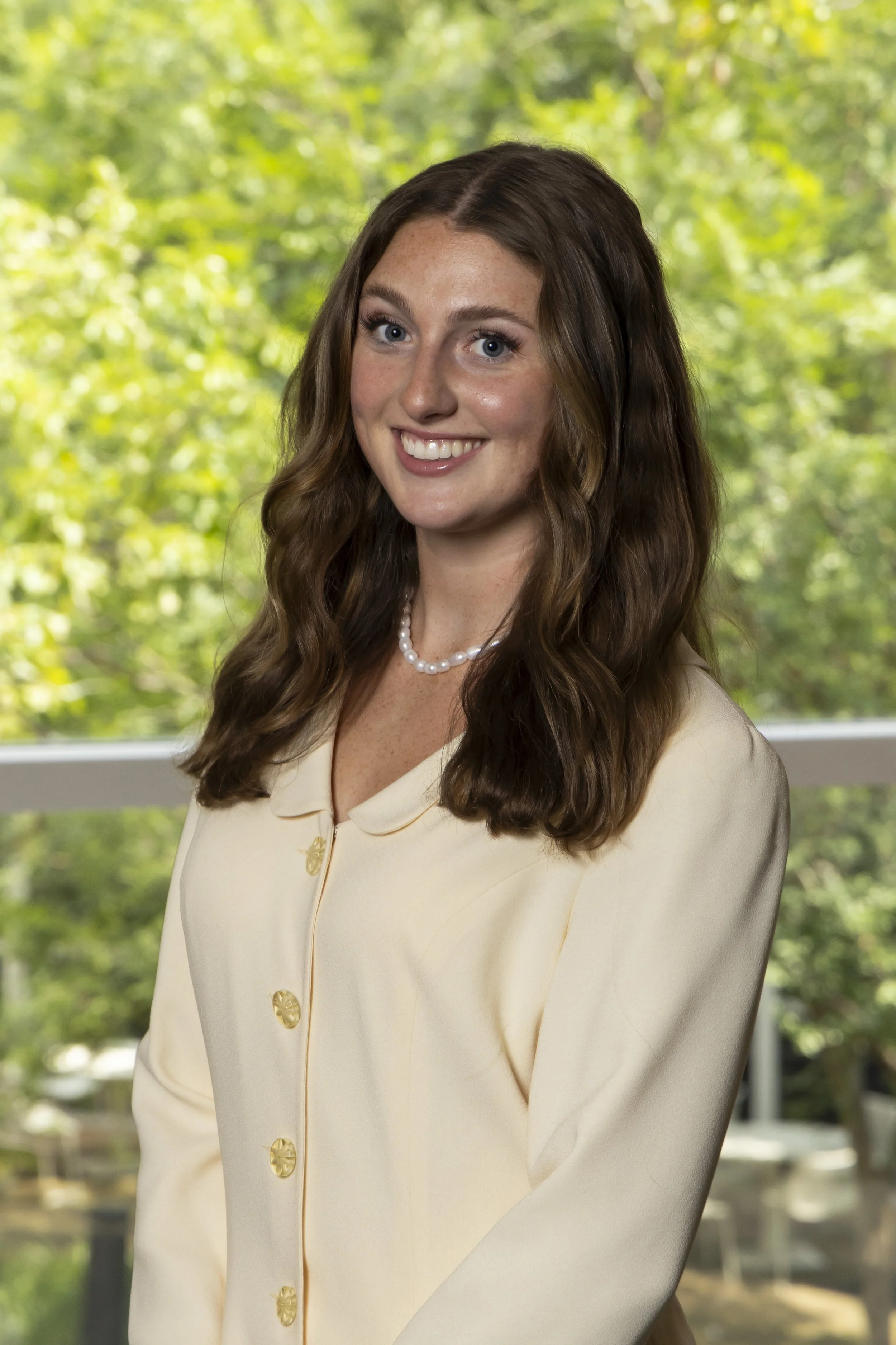 A young woman with long wavy brown hair, wearing a cream-colored blouse with gold buttons and a pearl necklace, smiling in front of a window with green trees outside.