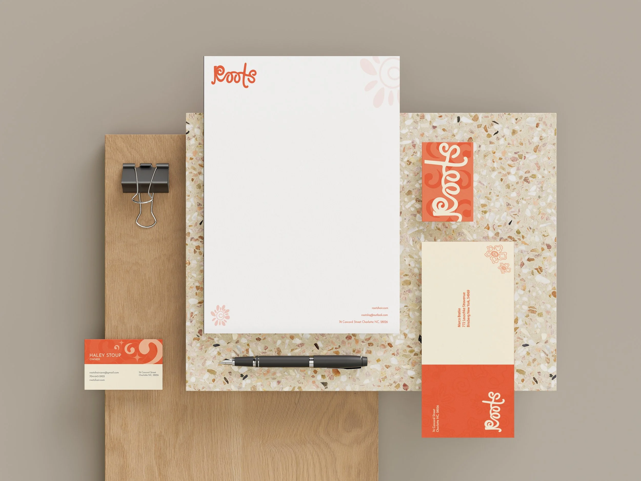 Flat lay of branded stationery featuring "Roots" logo. Includes letterhead, business cards, and an envelope on a wooden and terrazzo surface, with a paper clip and pen.