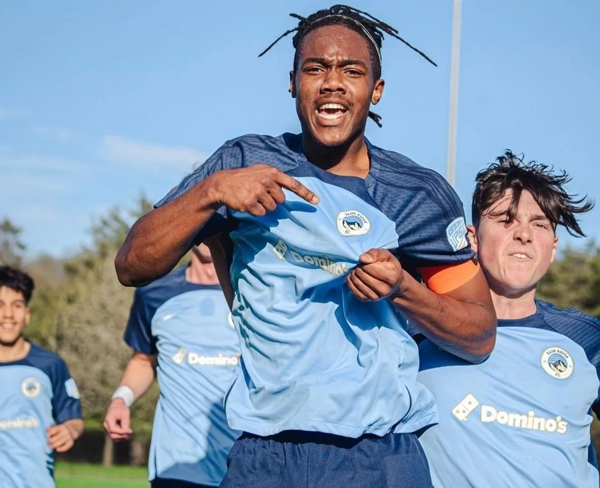 Group of soccer players in blue jerseys celebrating on a field, with one player pointing at the team logo.