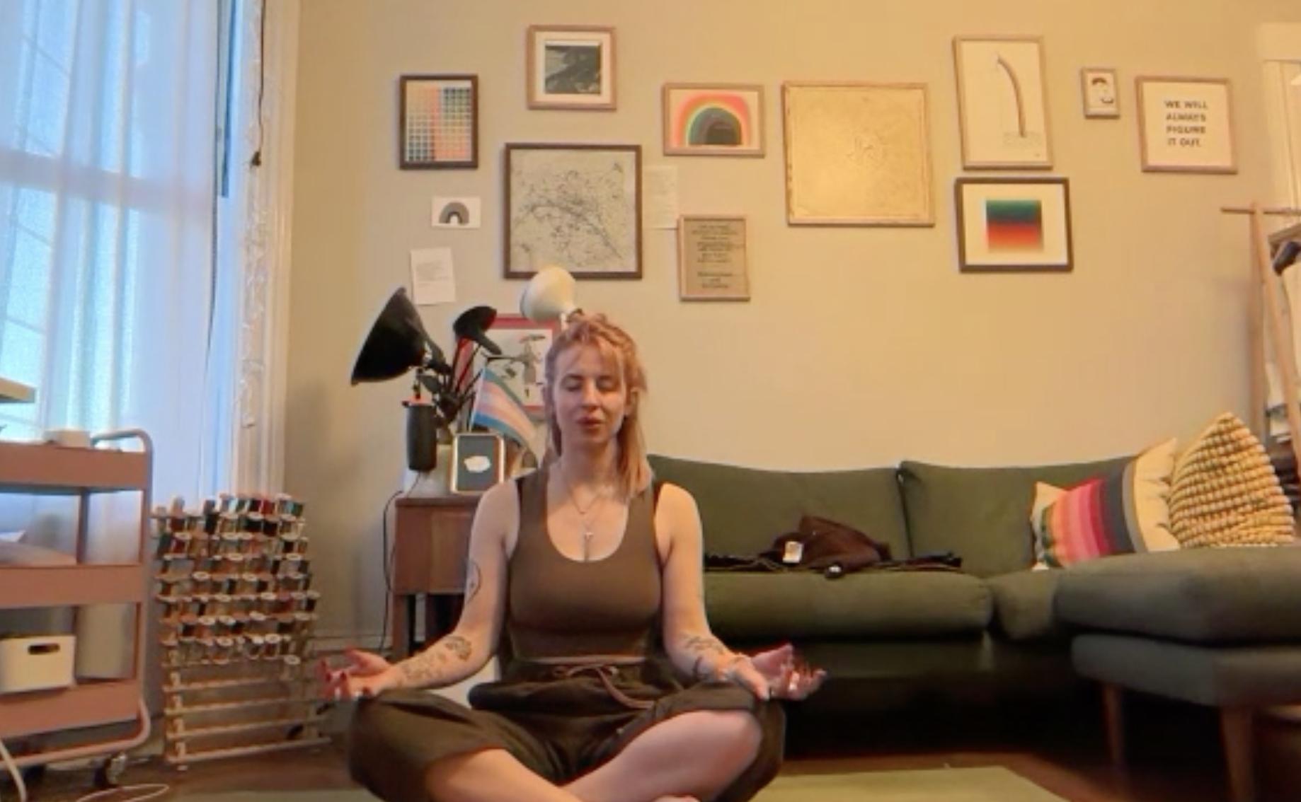 A woman with tattoos sitting cross-legged and meditating in a living room, with a green couch, colorful pillows, and a gallery wall of framed artwork behind her.