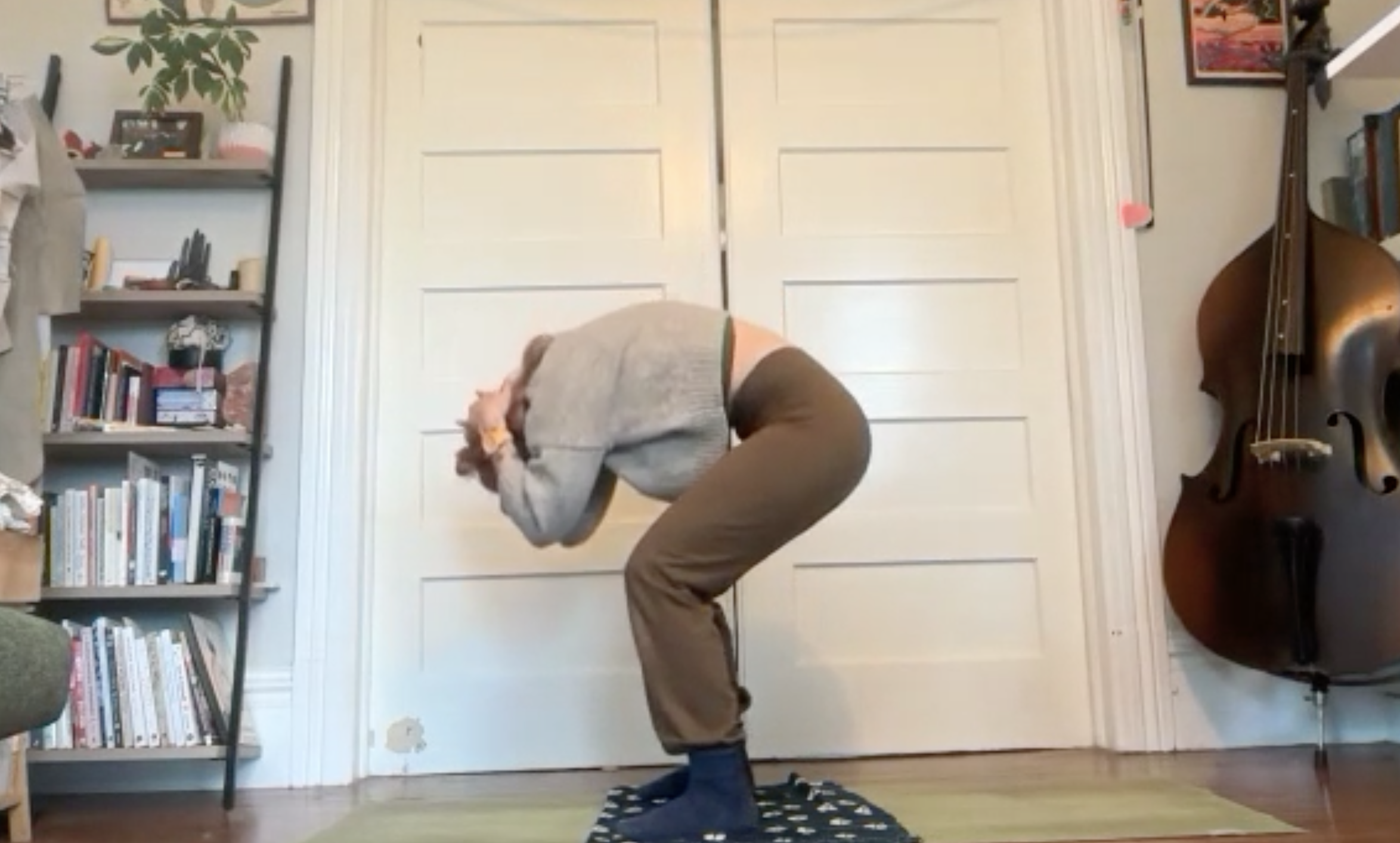 Person bending forward with hands over face, standing on a small rug in a room with bookshelves, a double door, and a guitar.