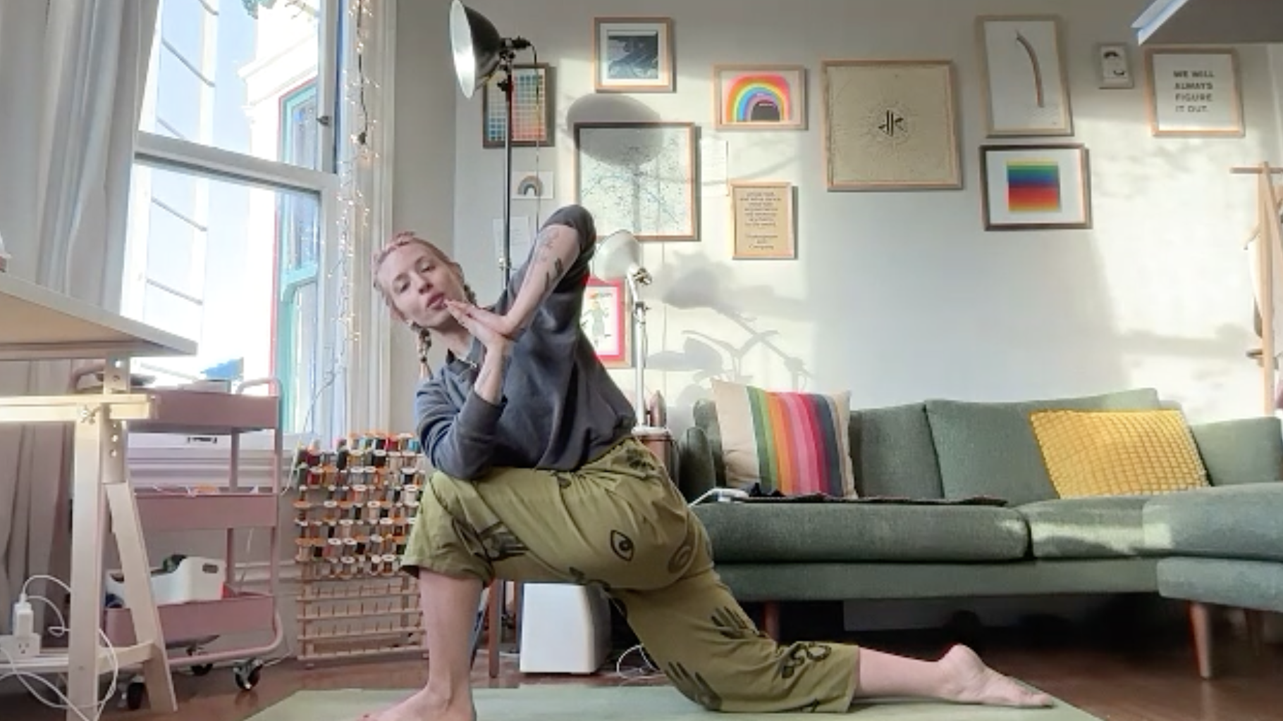 A woman practicing yoga in a cozy living room with artwork on the wall, a green sofa with colorful pillows, and sunlight streaming through a large window.