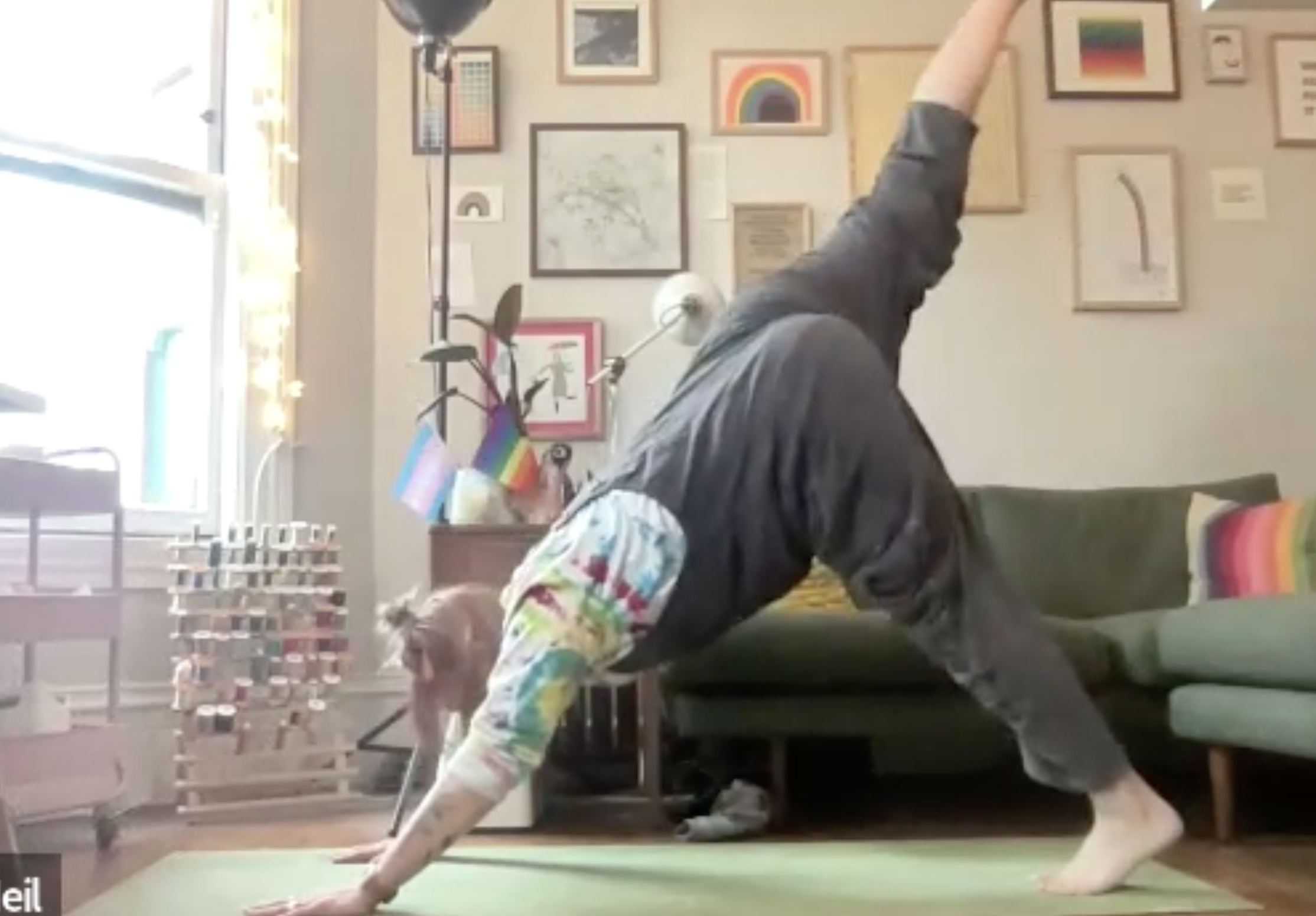 Person in athletic wear doing a yoga pose or stretch on a green yoga mat in front of a wall decorated with colorful framed artwork, rainbow-themed items, and a window with sunlight.