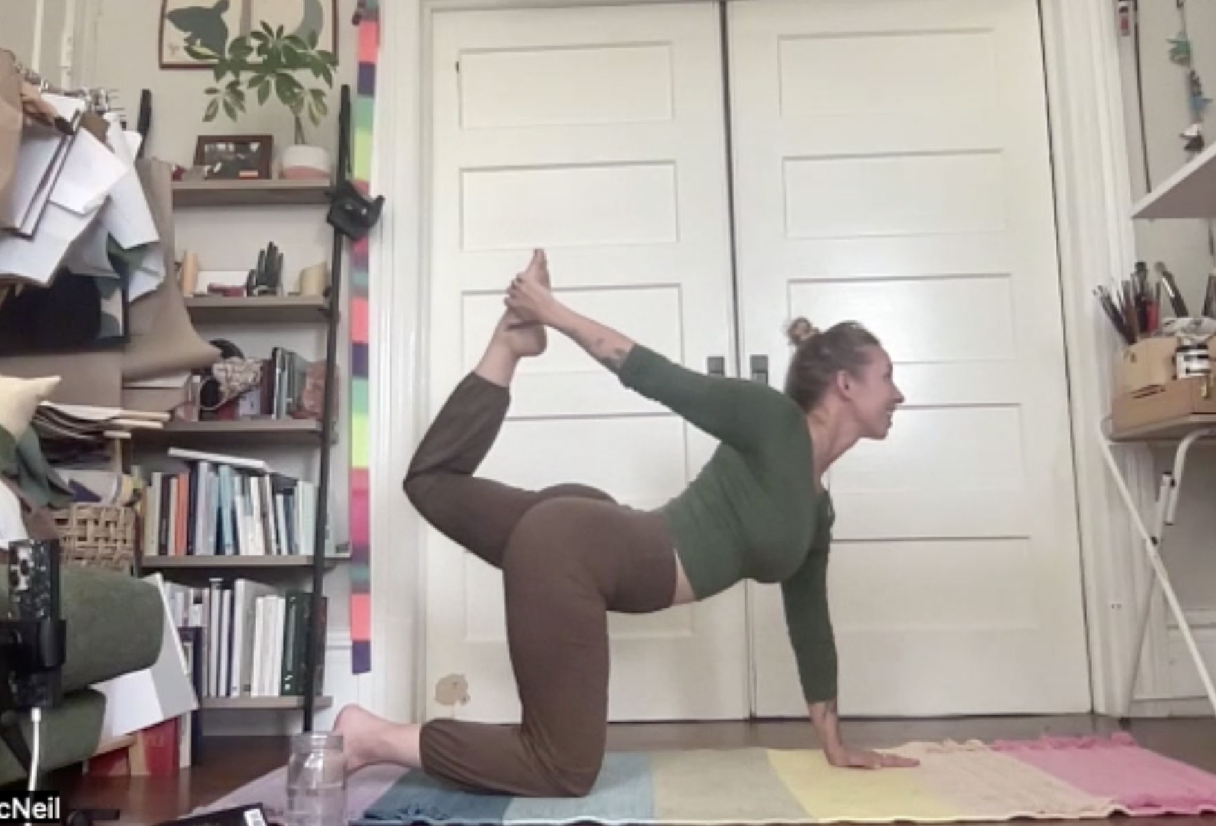 A woman practicing yoga in a room, balancing on her hands and knees with her back arched and one leg lifted, holding her ankle with her hand.