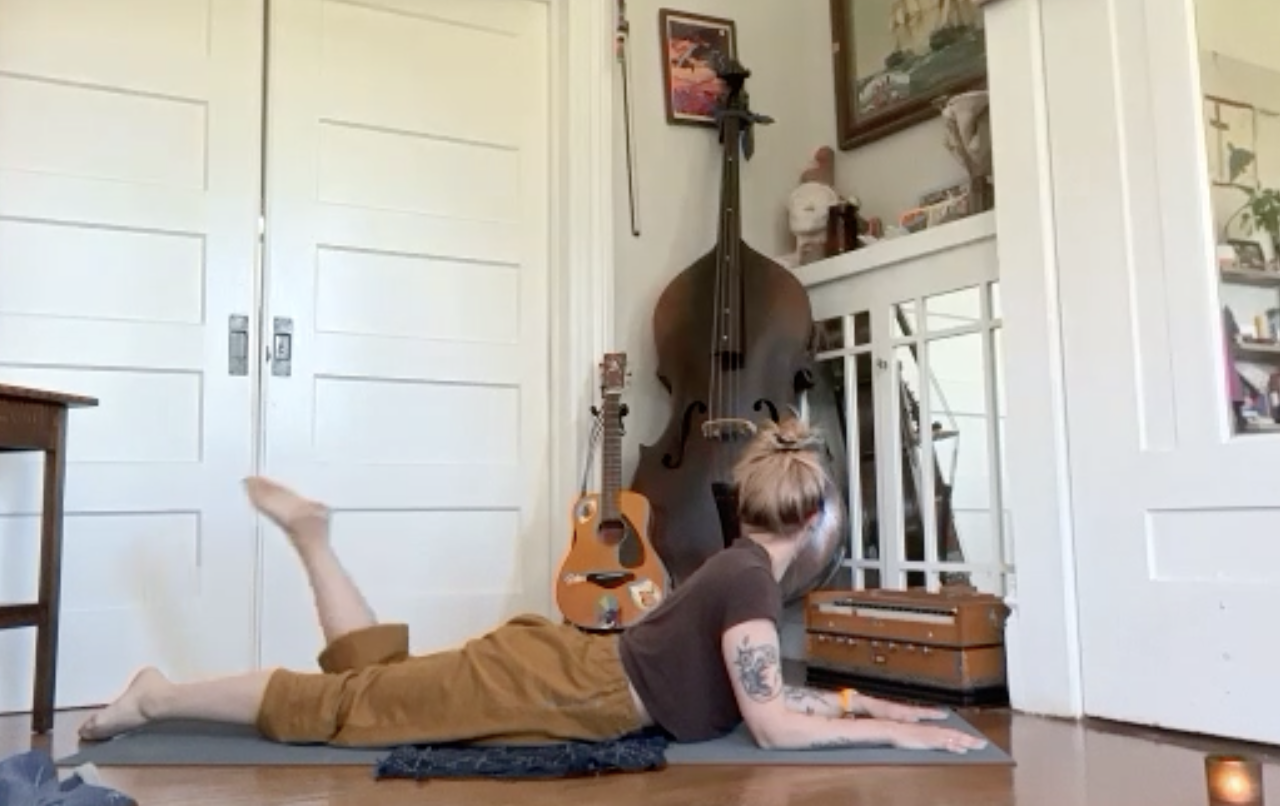 A person with tattooed arms lying on their stomach on a yoga mat, facing a large upright double bass in a room with guitars and artwork on the walls.