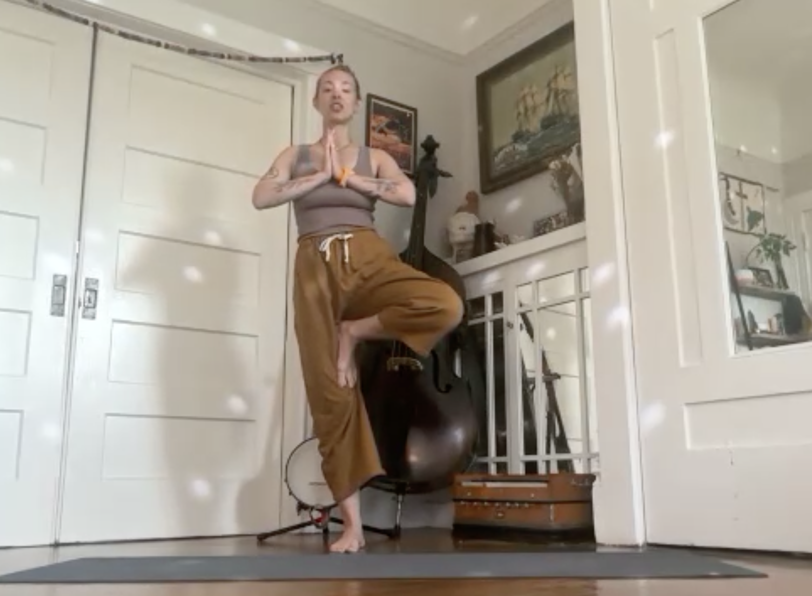 A woman practicing yoga indoors, standing on a yoga mat with one foot on her thigh in a tree pose, hands in prayer position, in a room with artwork, plants, and musical instruments.