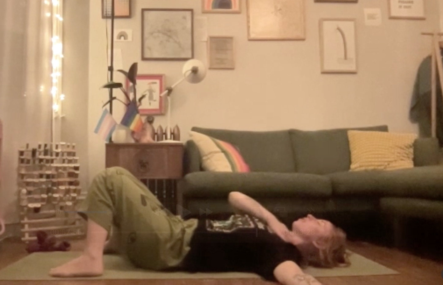 Person lying on their back on a yoga mat in a living room, stretching with one arm over their head, with a green couch and wall of framed pictures behind them.