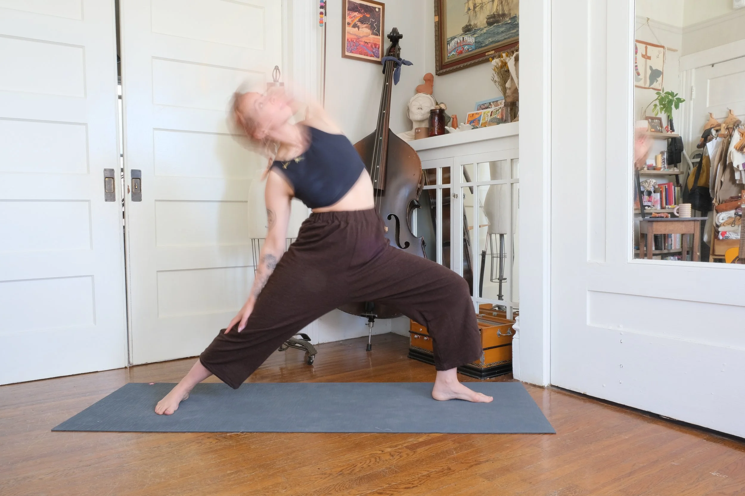 A person practicing yoga indoors, performing a wide-legged stance with one arm extended and the other hand on the floor. The room has a wooden floor, a gray yoga mat, and a white closet door, with various artworks, a double bass, and decorative items on shelves in the background.