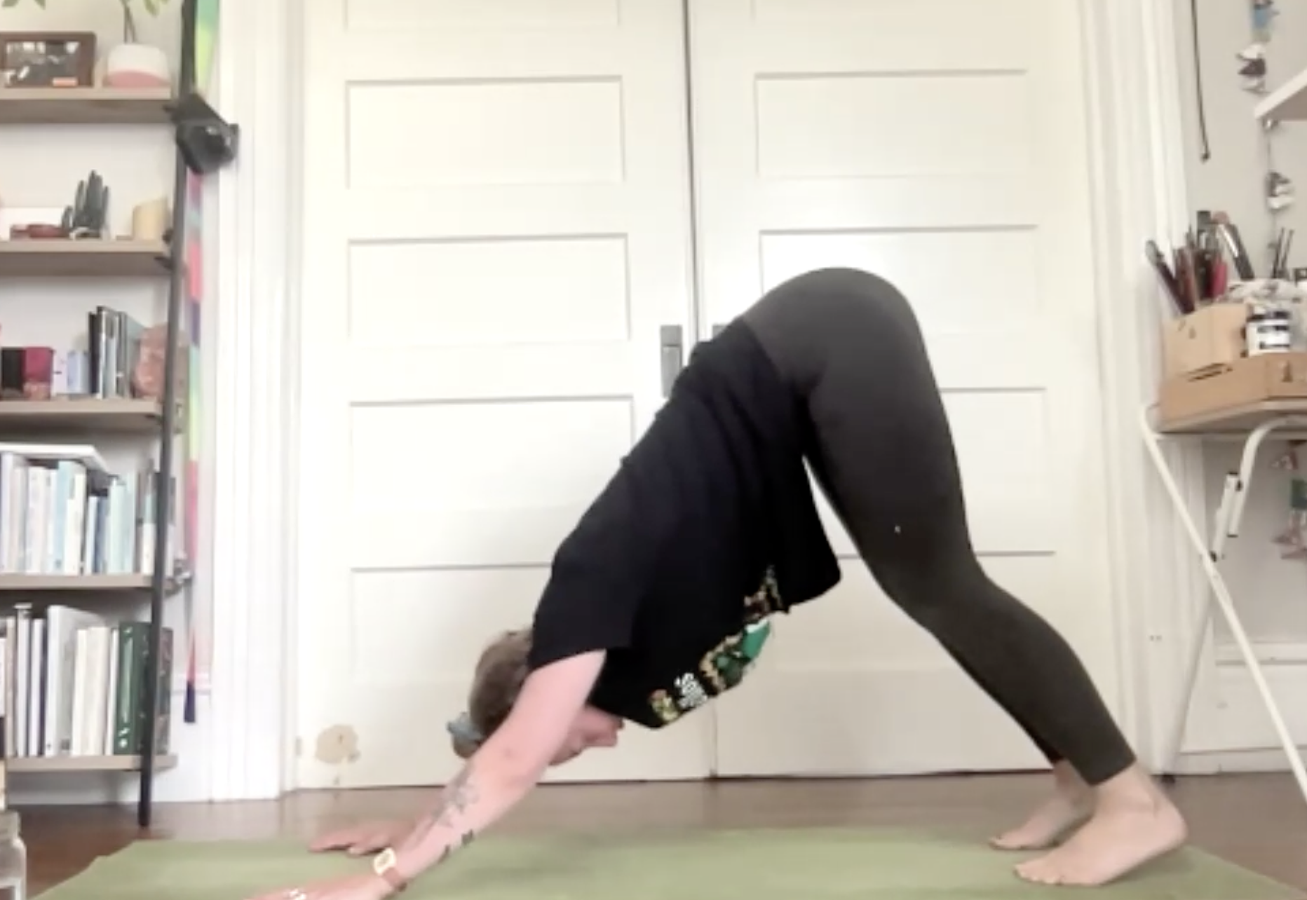 Person in black shirt and dark pants performing a downward dog yoga pose on a green mat in a room with shelves and closed white doors.