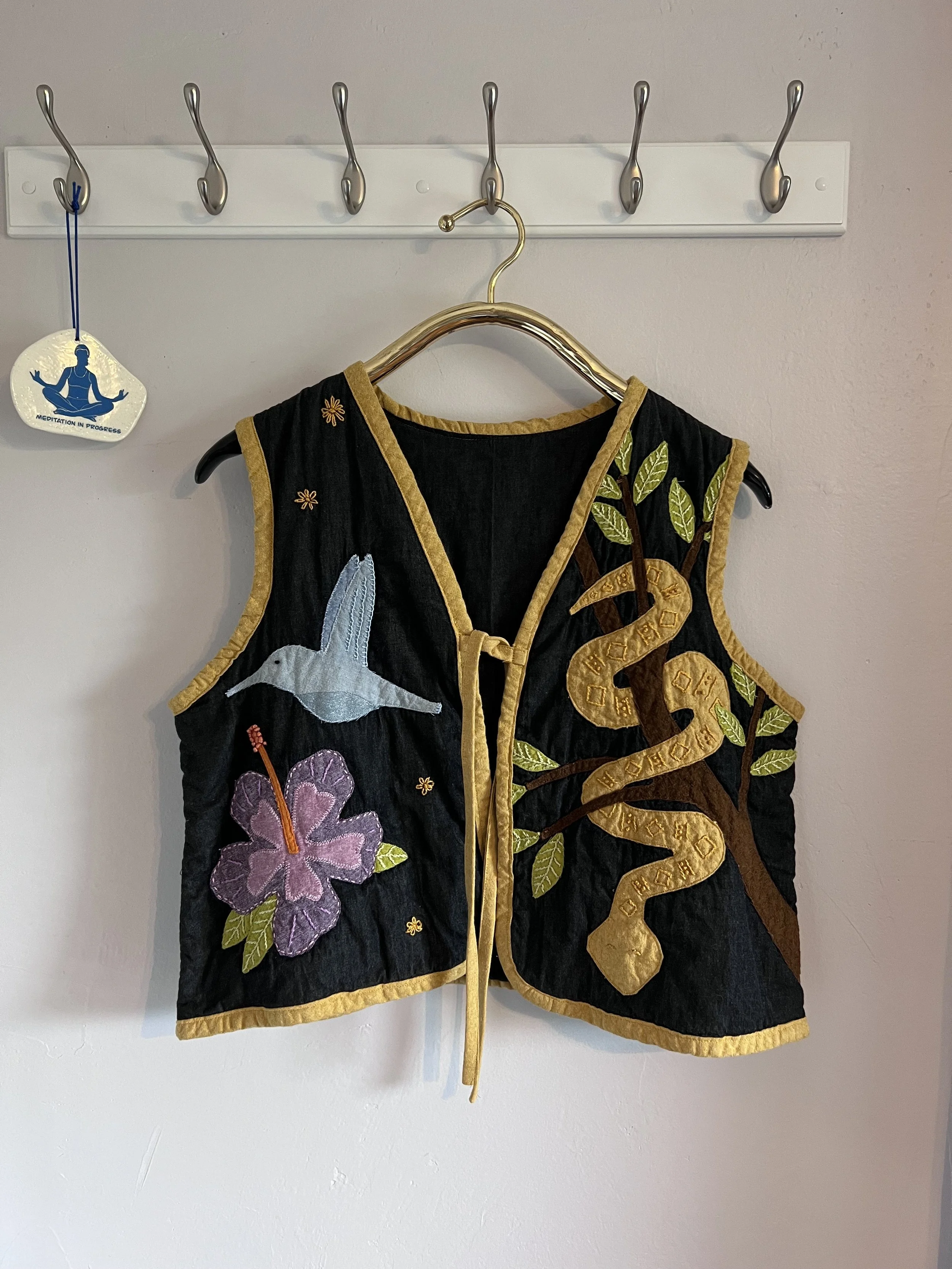 Black vest with embroidered hummingbird, purple flower, snake, and plant motifs, hanging on a wall hook with a meditation tag nearby.