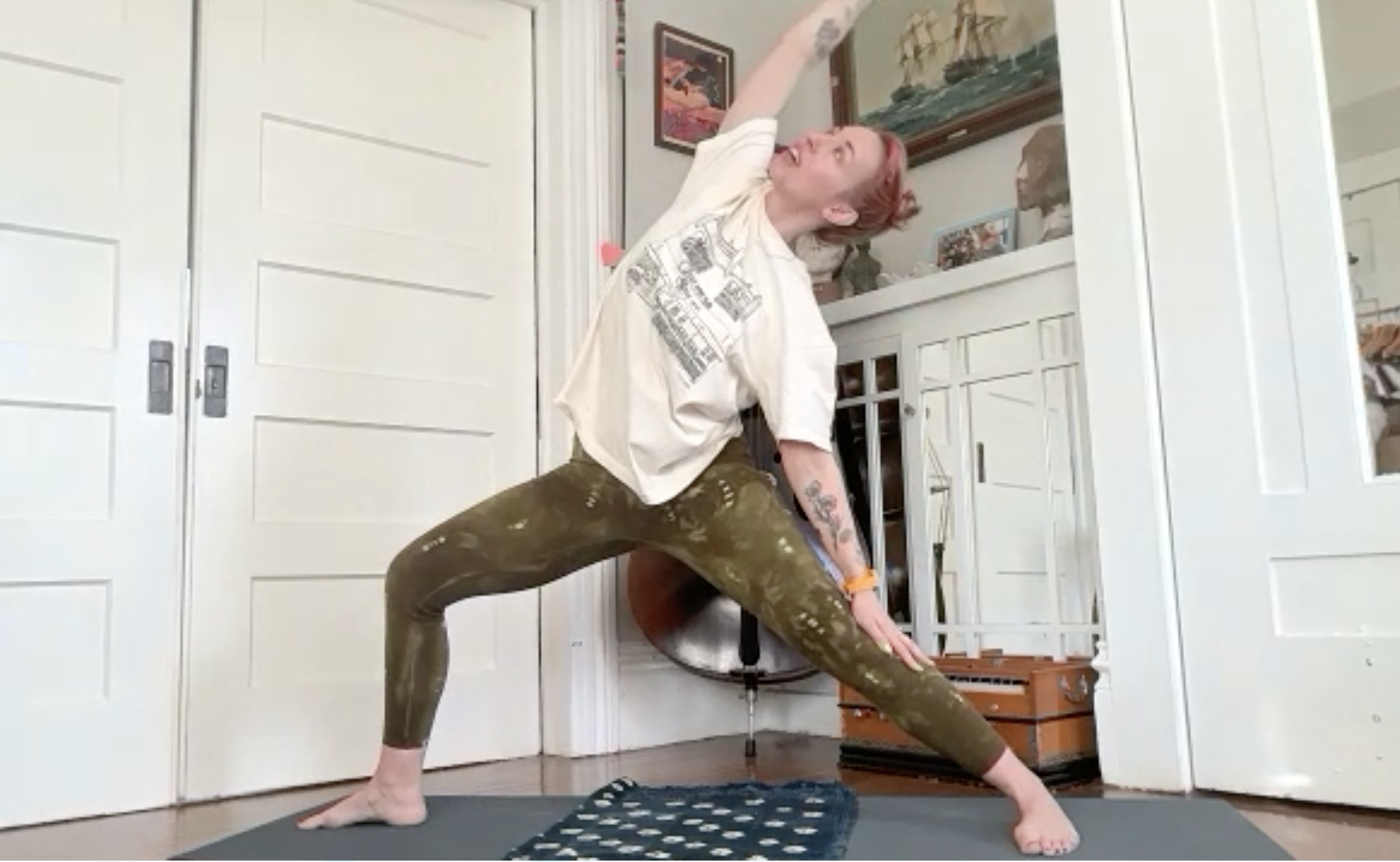 Person practicing yoga indoors, performing a pose with one leg bent and one arm reaching overhead, in front of a white closet door and houseplants.