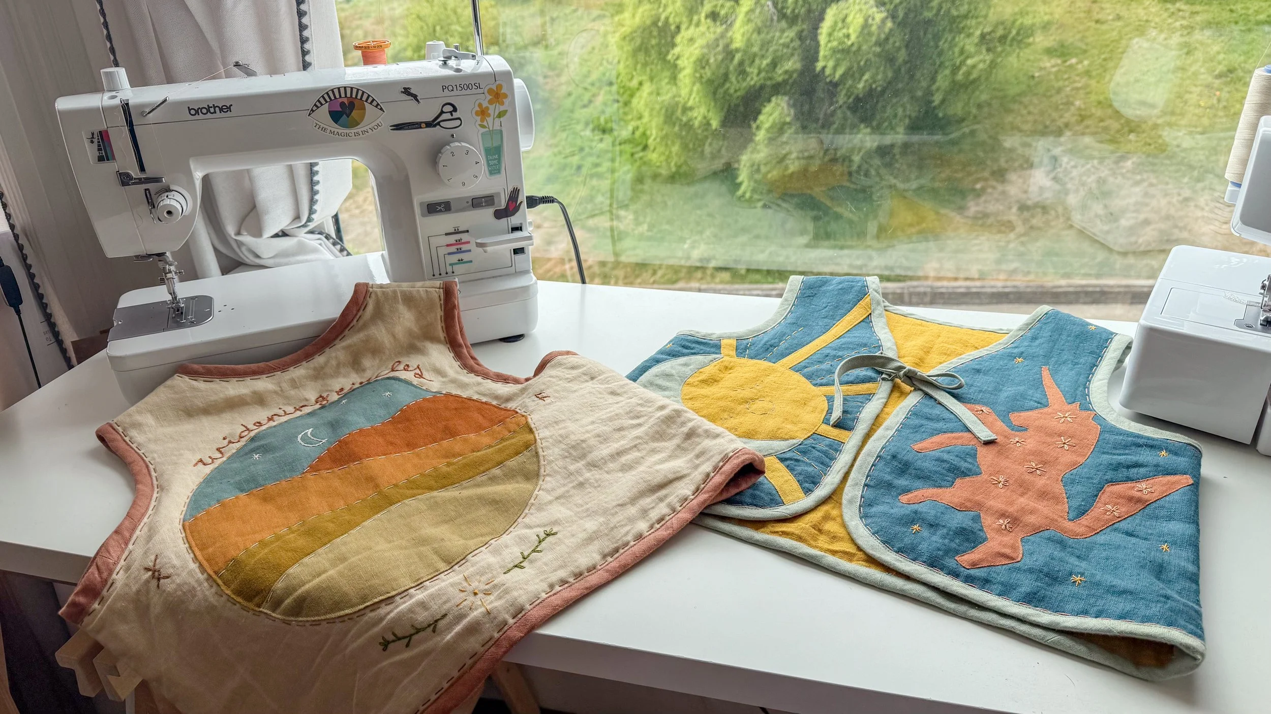A sewing machine on a white table with three colorful patchwork bibs featuring a cactus with a moon, a sun, and a pink fox; window with greenery outside in the background.