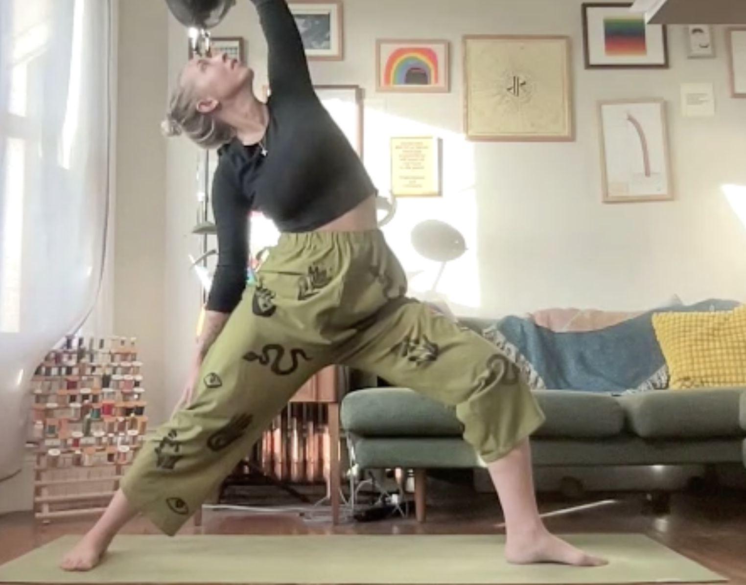 Person practicing yoga in a living room, in a wide-legged forward bend pose with one hand touching the floor and the other reaching upward, wearing a black long-sleeve top and tan pants with black patterns.