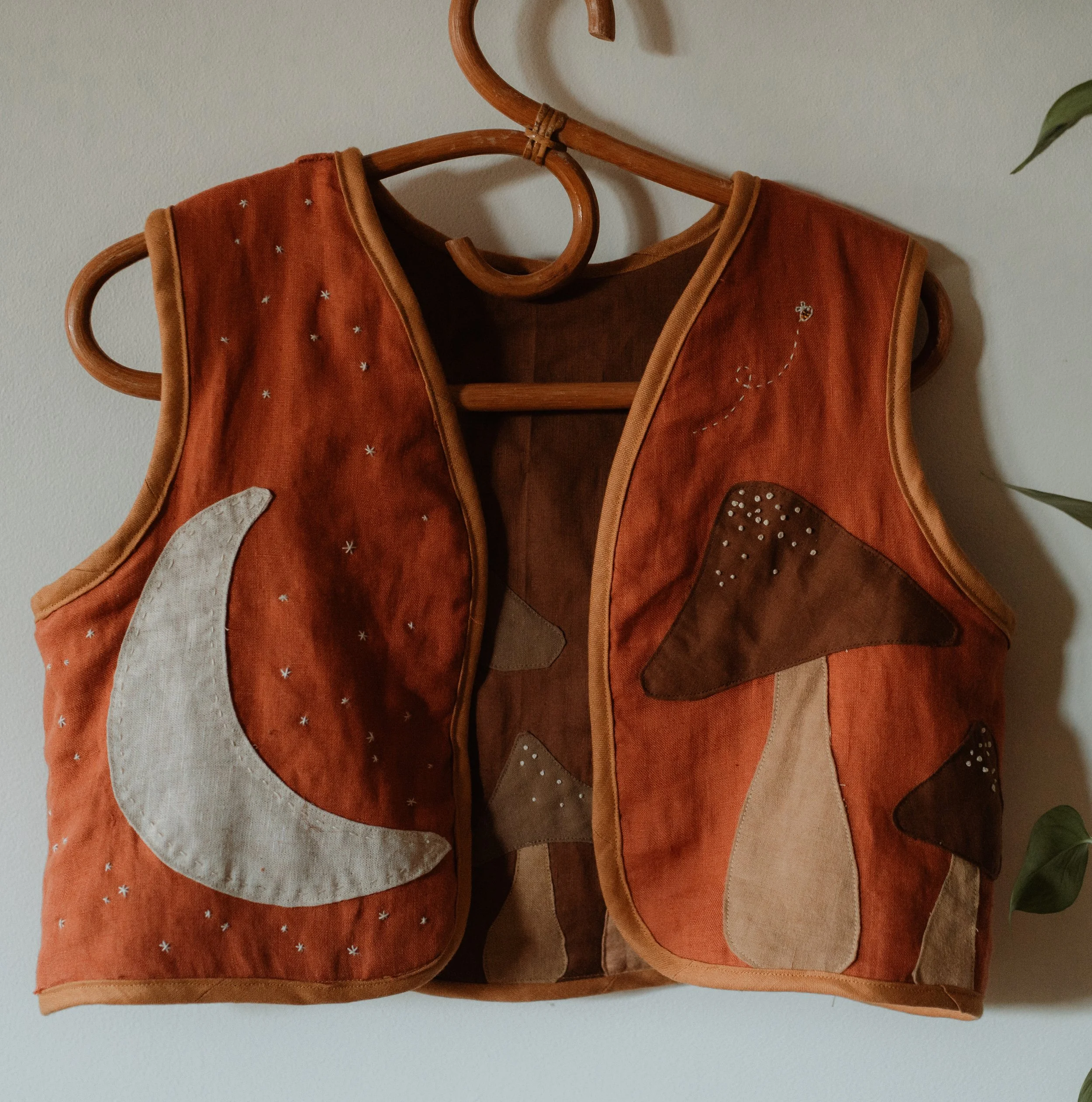 A sleeveless quilted vest with celestial applique, featuring a moon on the left side and a star and planets on the right side, hanging on a wooden hanger against a neutral wall.