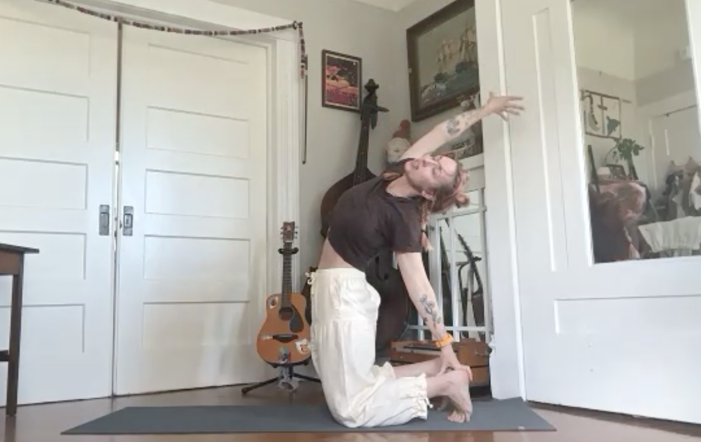A person with tattoos and glasses in a yoga pose on a mat in a room with musical instruments and artwork.