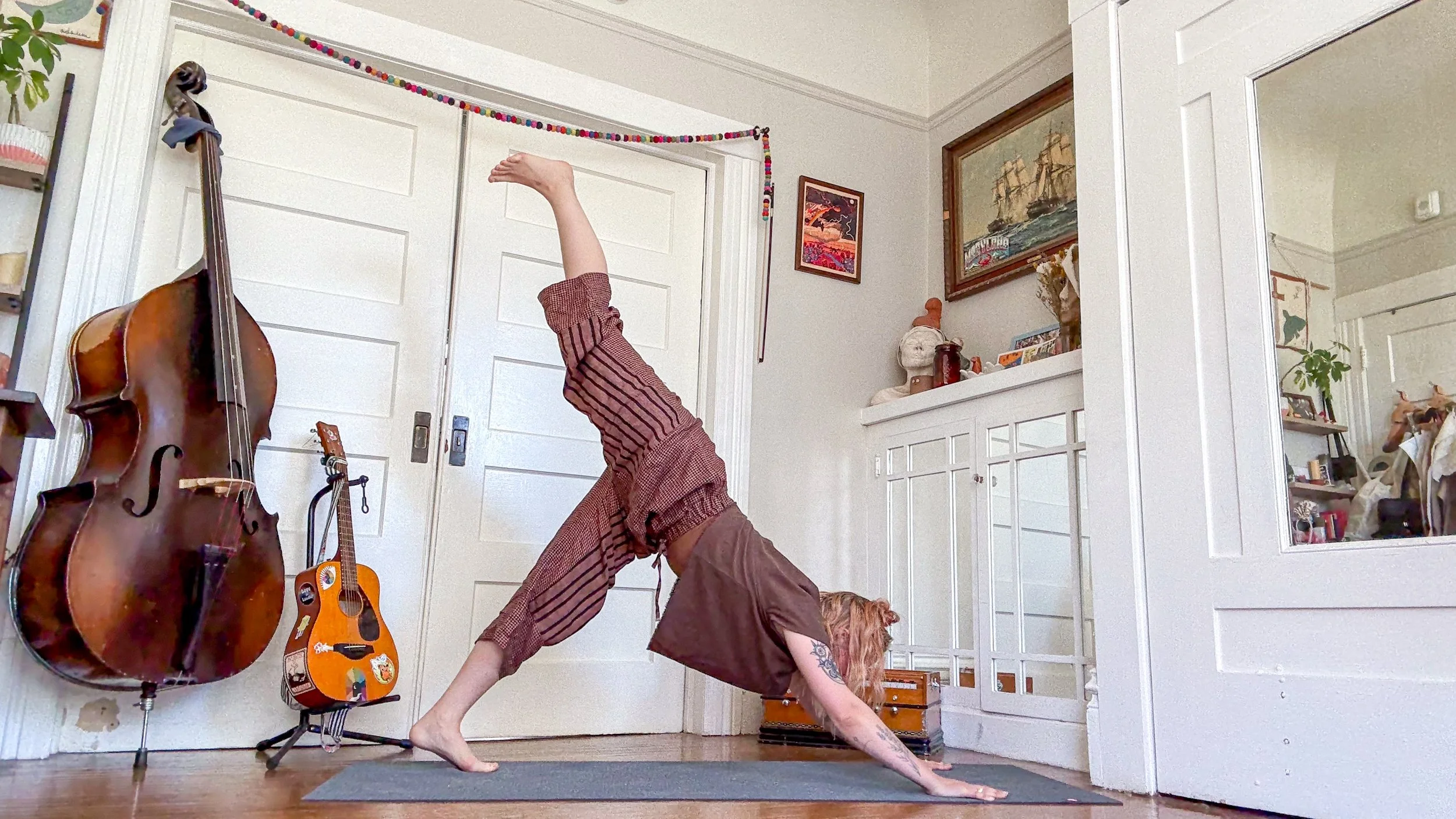 A person practicing yoga in a home with musical instruments, including a large acoustic guitar and a smaller guitar, on the floor. The person is in a pose with one hand on the floor, one leg extended straight up, and the other leg bent. The room has white walls with art and decorations, and there is a mirror reflecting part of the room.