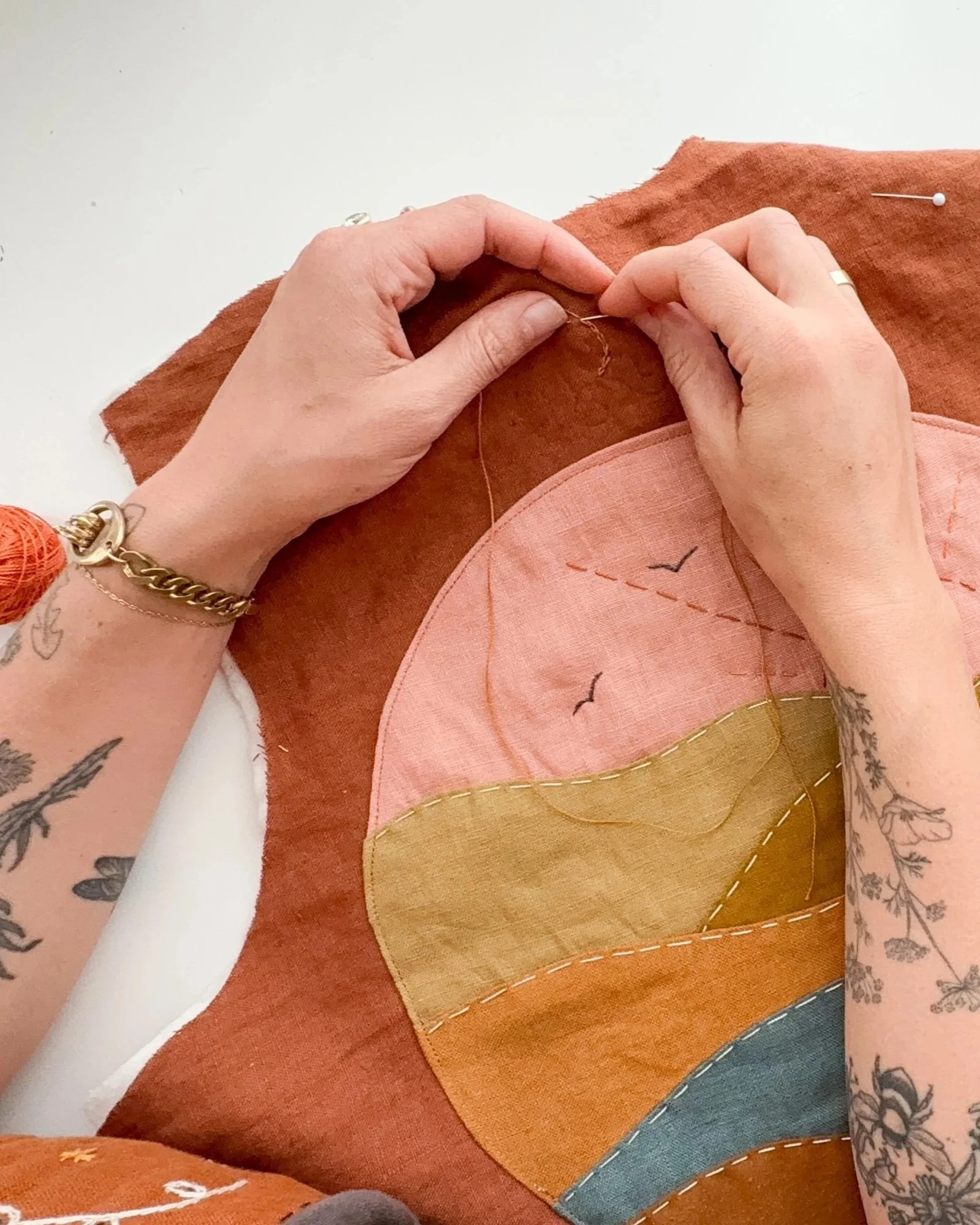 Person sewing colorful fabric with a needle and thread, with tattoos on their arms and wearing jewelry.