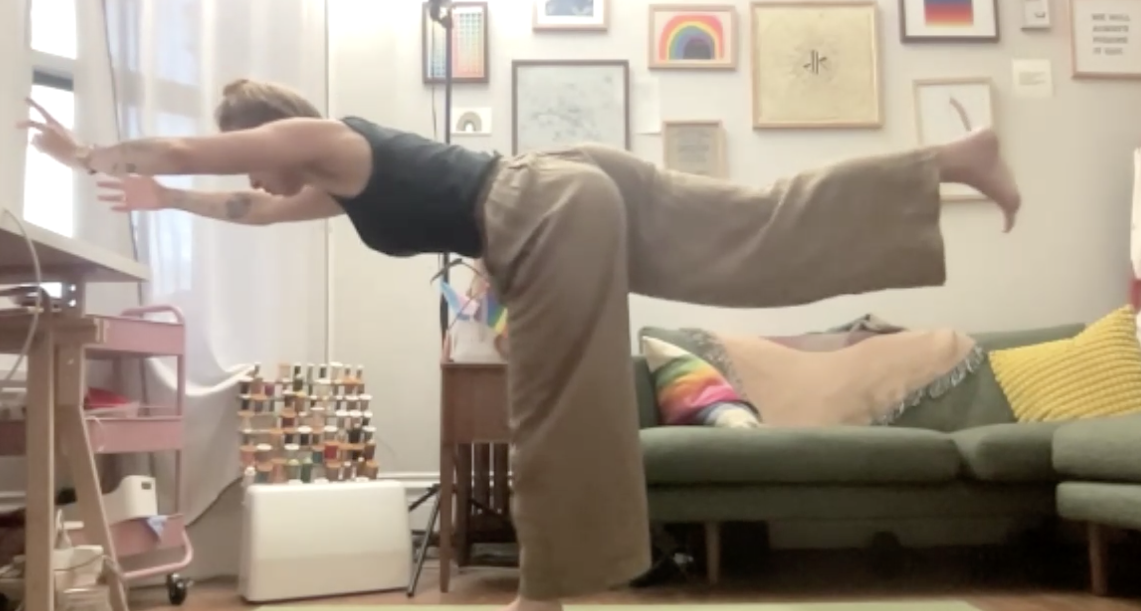 A person is performing a yoga pose in a living room, leaning forward with arms extended and head down, wearing a black shirt and loose beige pants. The room has colorful framed artwork on the wall, a green couch with multicolored pillows, a small pink table, and various toys and decorations around.