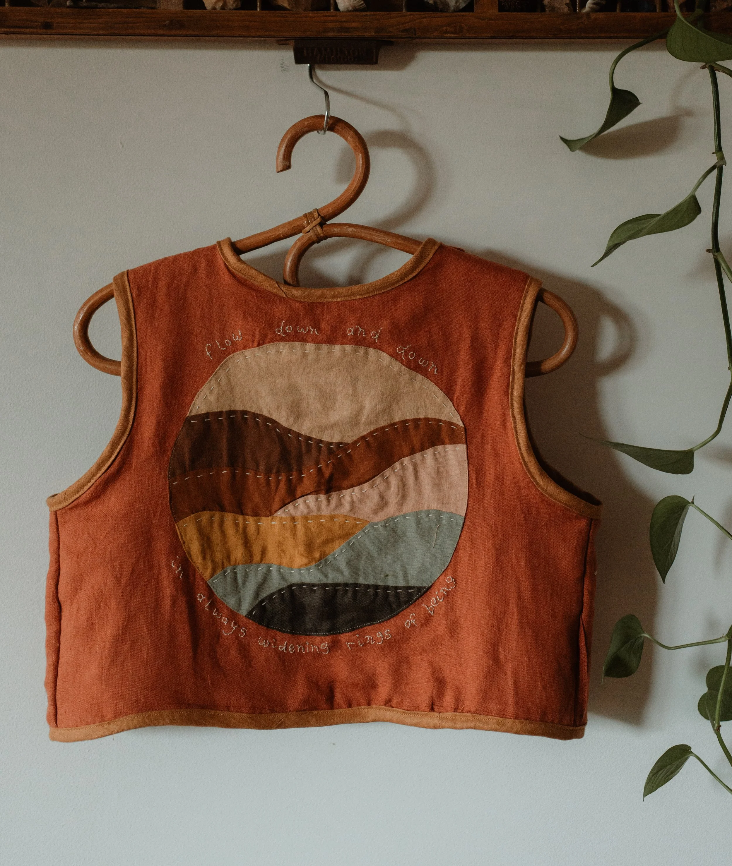 A sleeveless, rust-colored vest with stitched artwork of a circle containing a landscape of rolling hills in earth tones, and embroidered text that reads 'flow down and down as always winding rings or loops.'