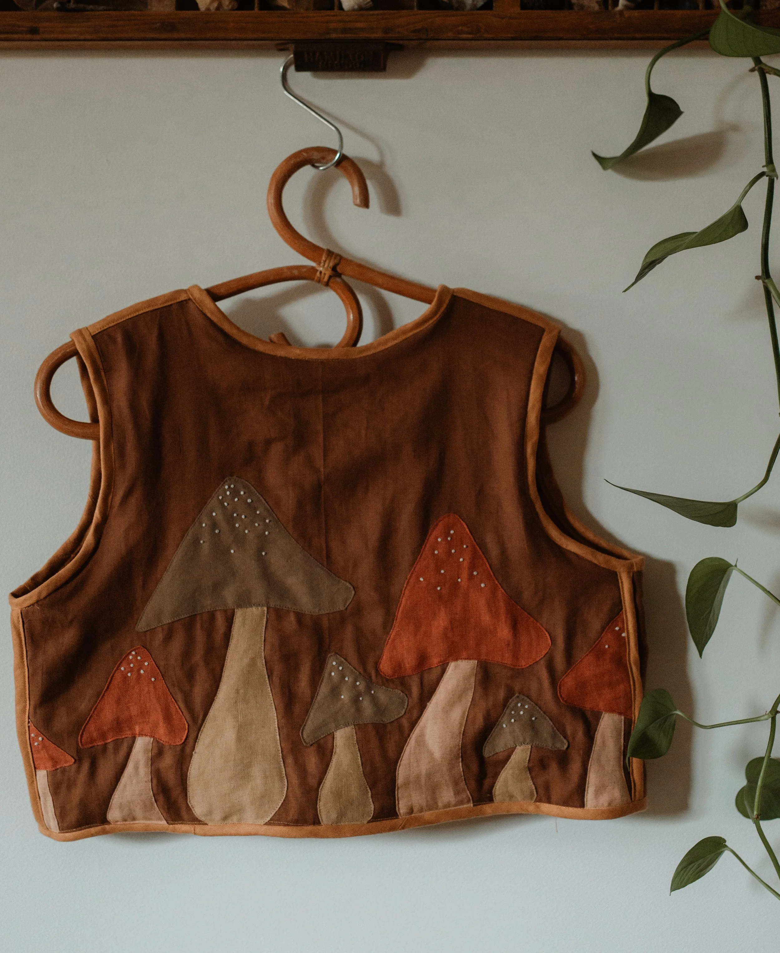 Leather vest with mushroom appliqué design hanging on a wooden hanger against a white wall.