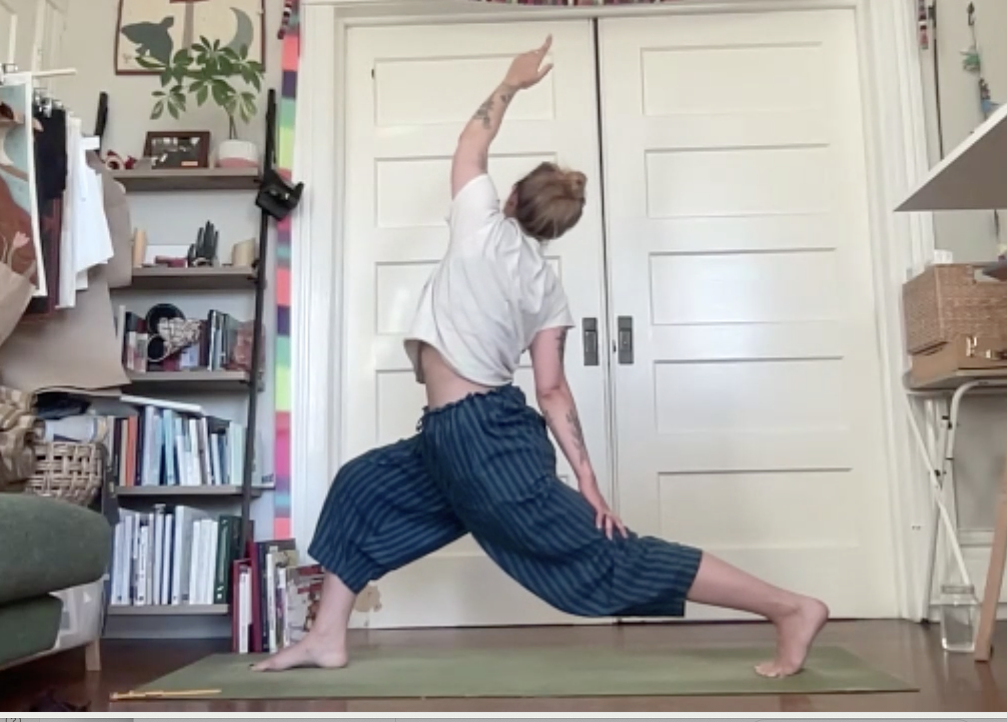 A person performing a yoga pose indoors, with one hand on the ground and the other arm raised, in front of a white double door and bookshelves filled with books and decor.