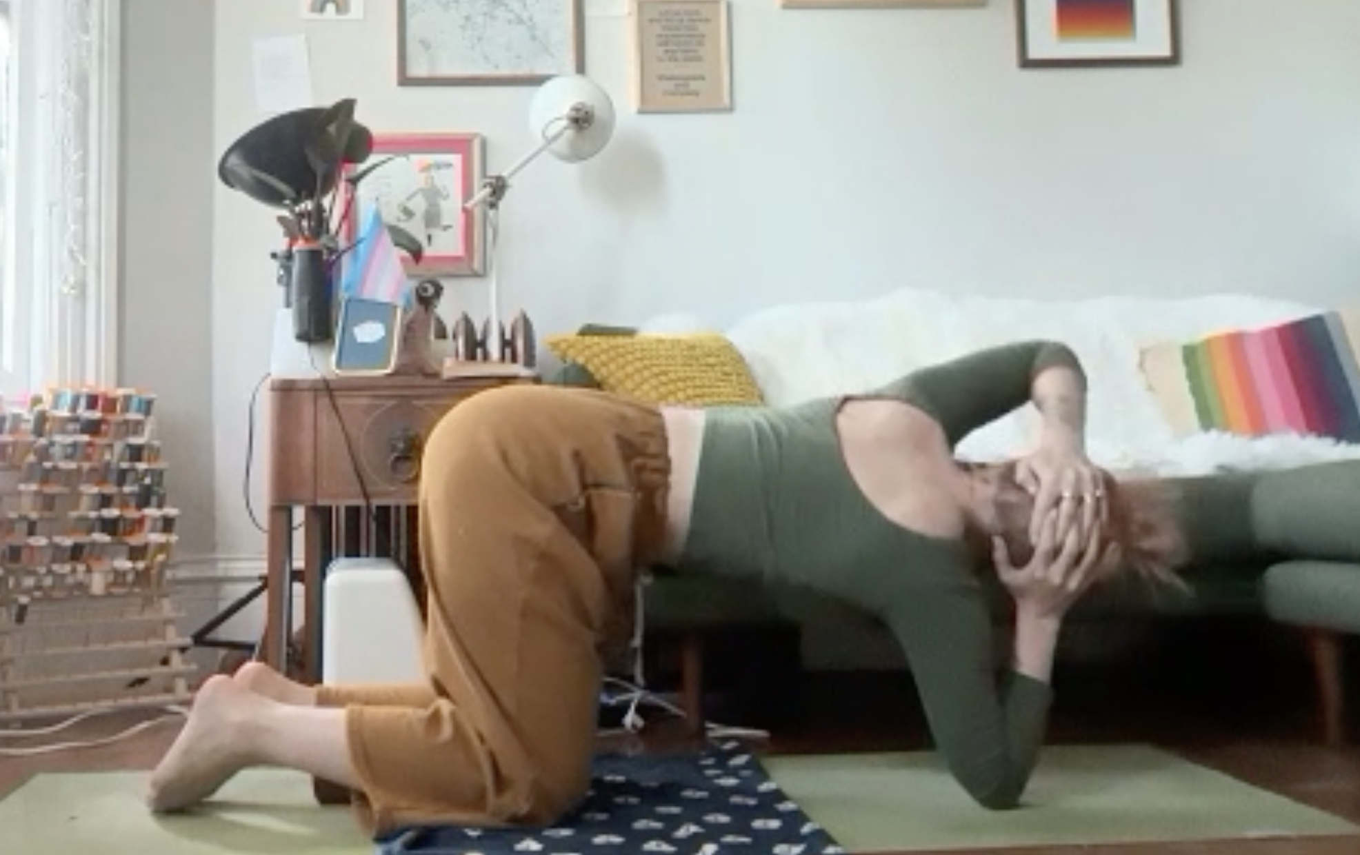 Person practicing yoga or stretching on a green mat in a living room, with one knee and their hand on the ground, head tilted down, surrounded by framed artwork and a bookshelf.