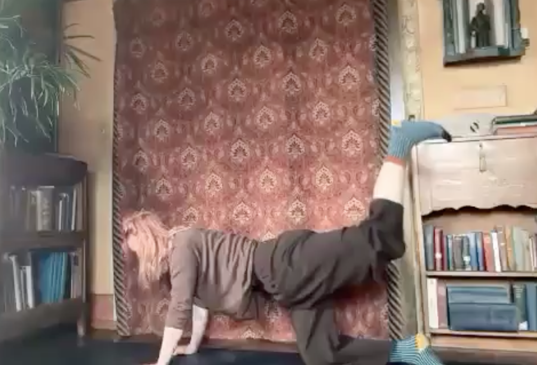 A person lying on a patterned red rug in a room with bookshelves on both sides, a yellow wall with a mirror, and a small plant in the corner. The person is wearing a gray hoodie, striped socks, and dark pants.