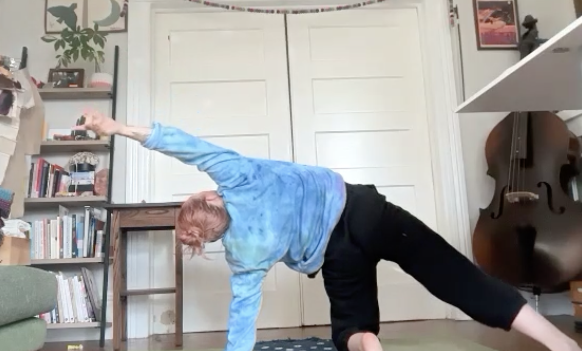 Person with red hair wearing a blue tie-dye shirt and dark pants performing a yoga pose in a living room with a white double door, a bookshelf, a framed picture, and a guitar.
