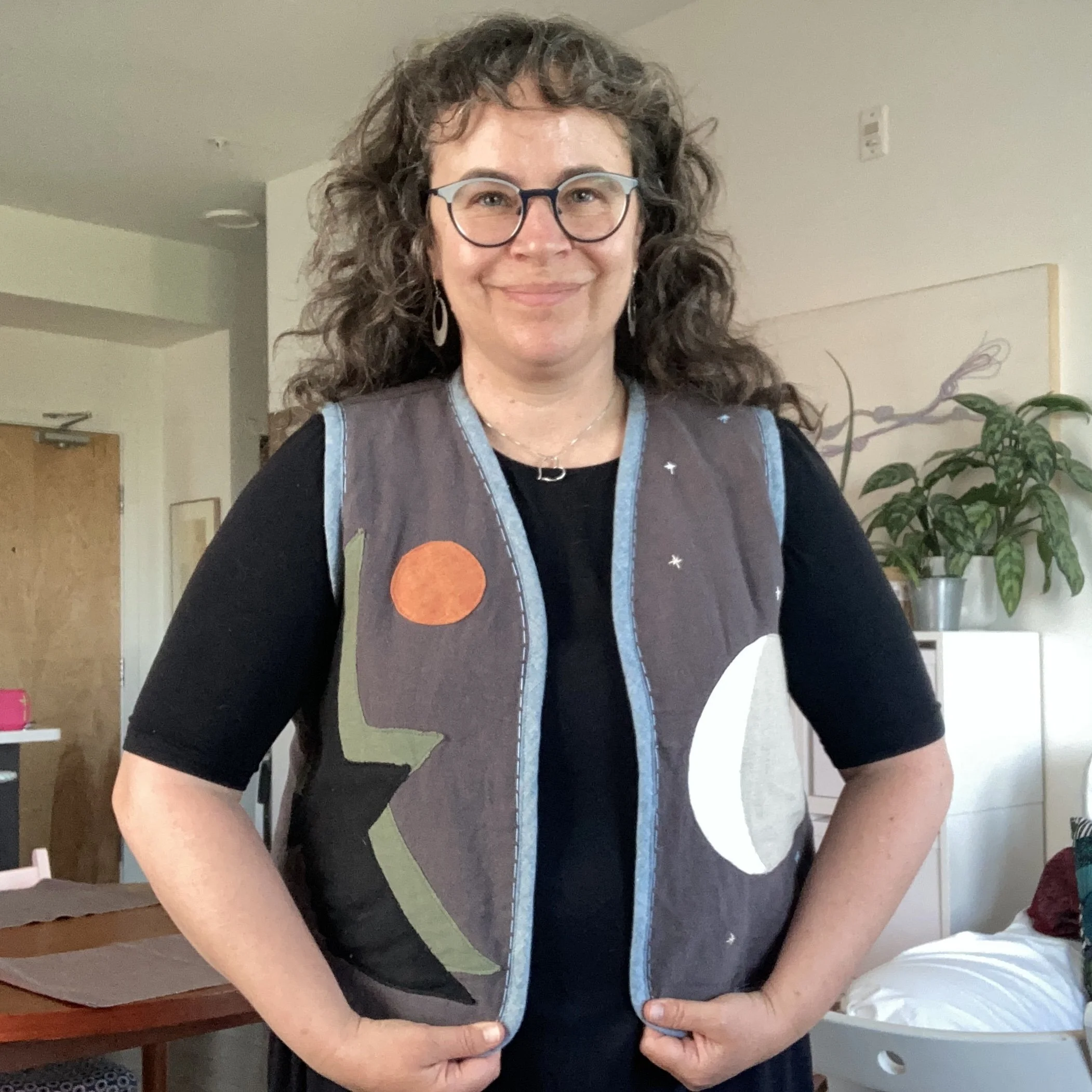 A woman with curly hair and glasses smiling, wearing a black shirt and a colorful vest featuring abstract moon and sun designs, standing in a room with plants and furniture.