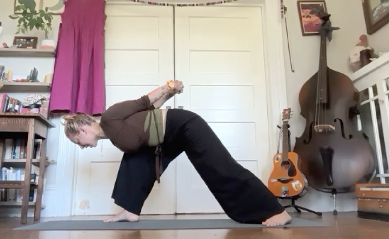 A woman practicing yoga indoors, in a pose with her hands on the floor and one leg extended backward, in a room with bookshelves, musical instruments including a double bass and an acoustic guitar, and artwork on the walls.