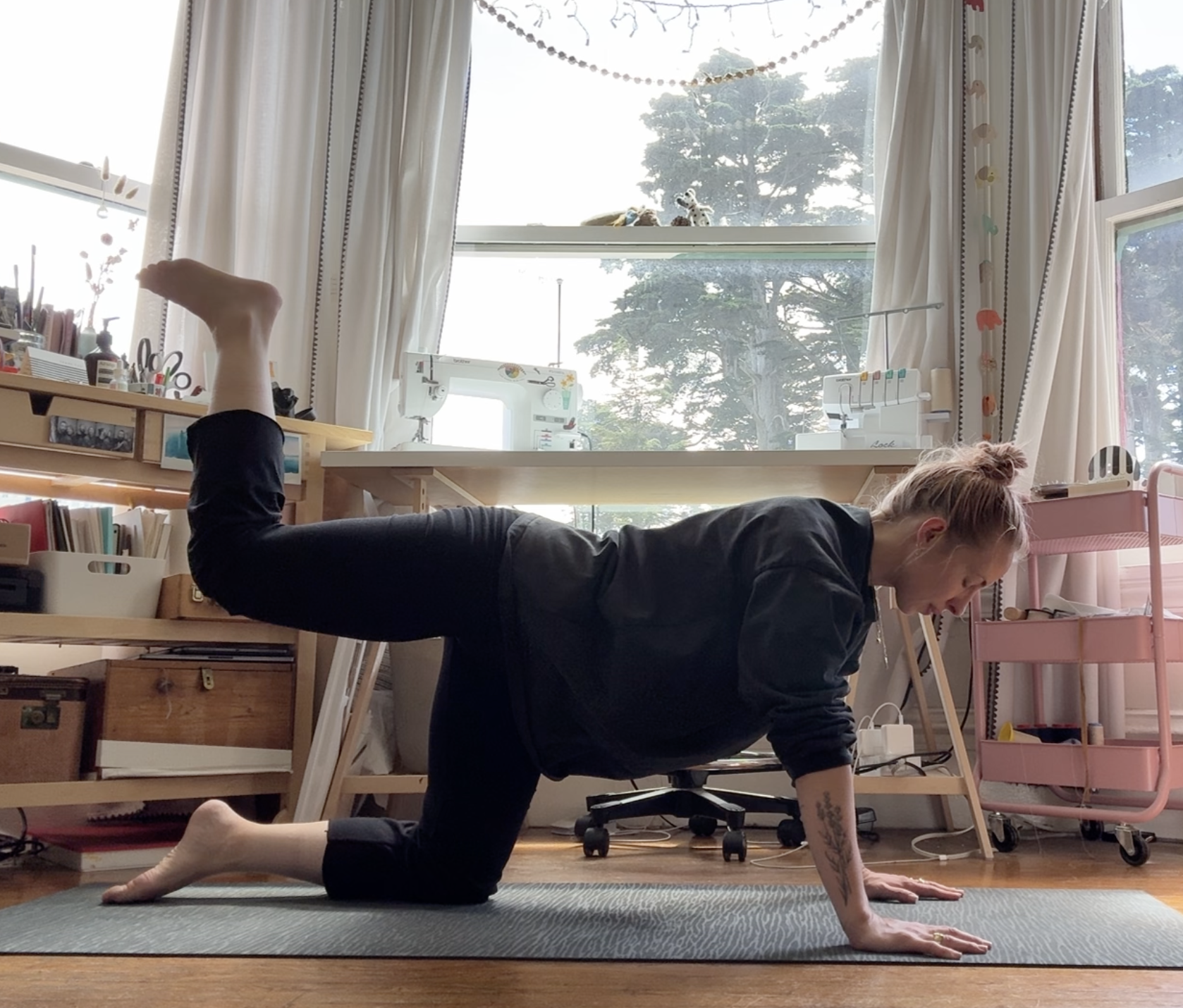 A woman doing a yoga pose in a well-lit room with large windows, books, and sewing machines, wearing a black long-sleeve top and black pants.