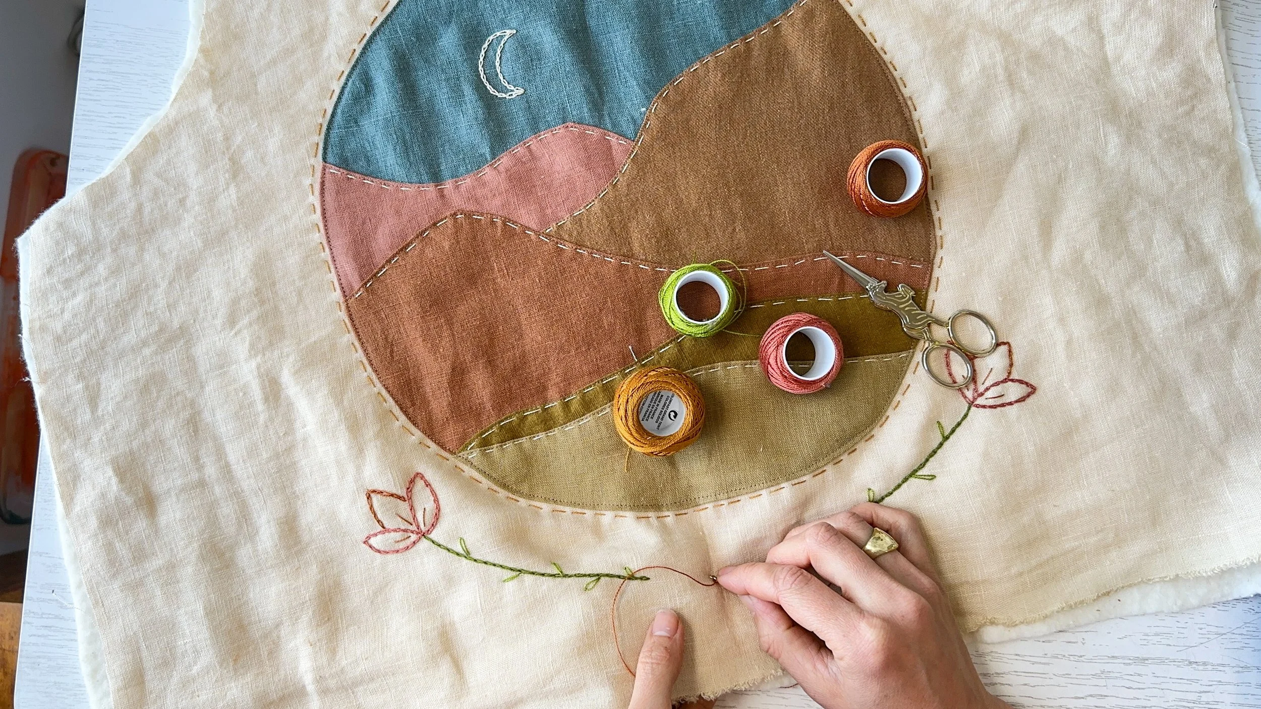 Embroidery in progress on a fabric with a landscape scene of colorful mountains, a crescent moon, and flowers. Several spools of thread in orange, pink, green, and yellow are present, along with a needle, scissors, and a hand holding the fabric.