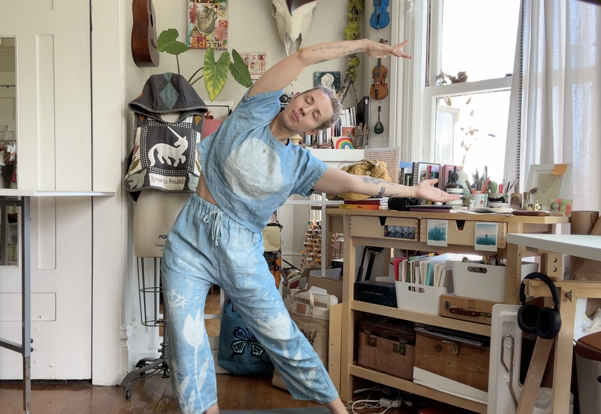 A person with curly blonde hair doing yoga in a cozy, art-filled room. They are barefoot, wearing a tie-dye or watercolor shirt and matching pants, with arms extended in a stretch. The room has musical instruments, books, plants, and decorative art pieces, creating an artistic and casual atmosphere.