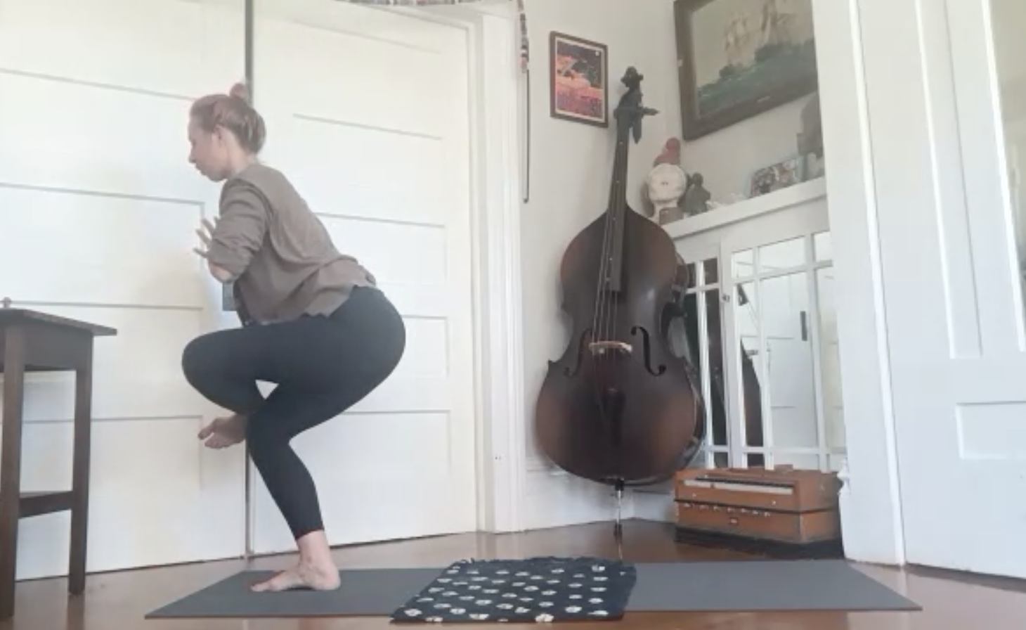 A woman practicing yoga in a room, balancing on her right foot with her left knee bent and raised, holding her hands clasped in front of her chest. The room has a double bass, framed artwork on the wall, and a wooden box on the floor.