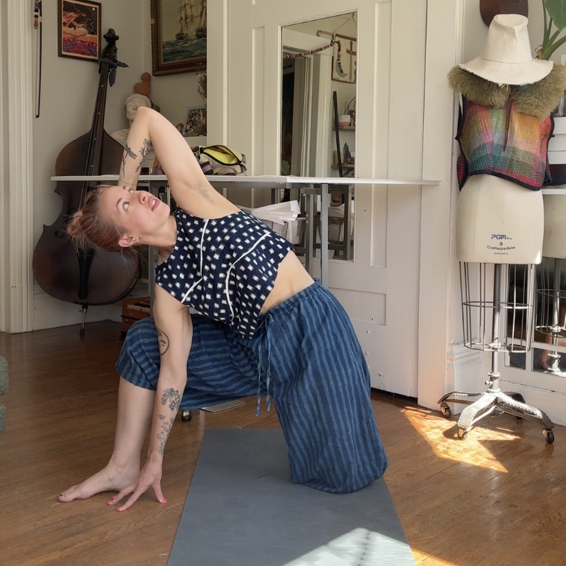 A woman practicing yoga indoors, in a pose where she balances on one hand and one knee, with the other arm extended overhead and head tilted back, on a yoga mat in a room with musical instruments and creative decor.