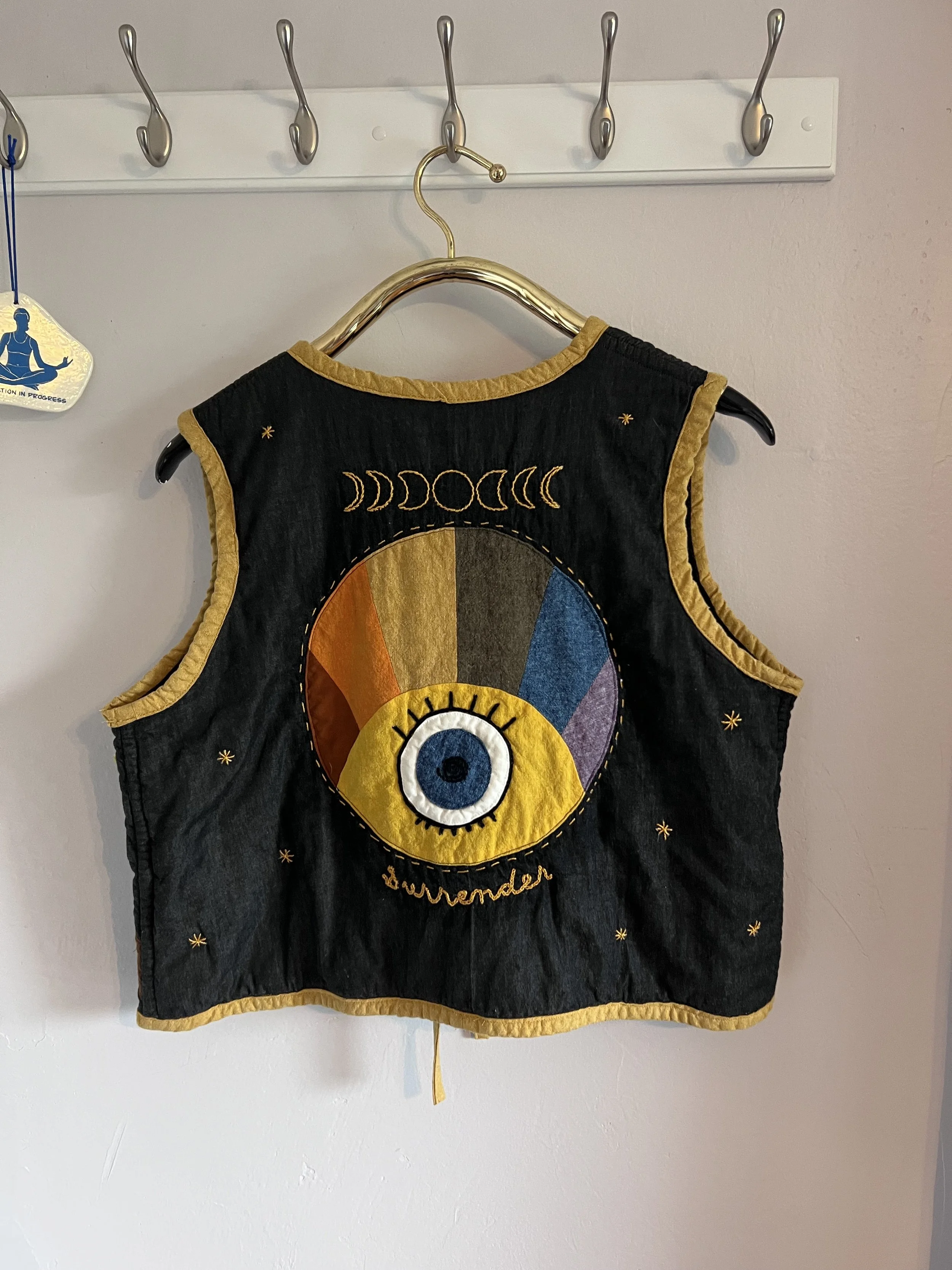 Black sleeveless vest with yellow trim, featuring an embroidered eye and a rainbow-colored arc, surrounded by moons and stars.