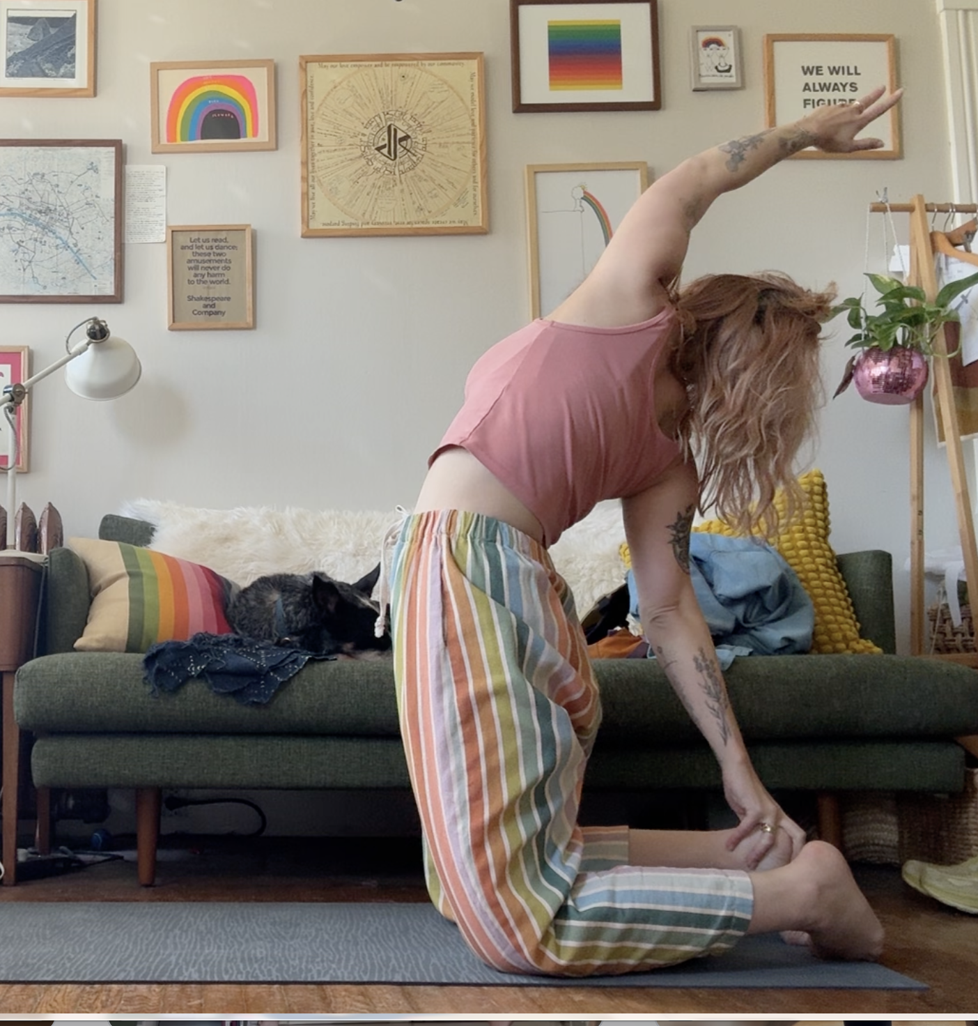 A woman practicing yoga on a mat in her living room, with a black and white cat on a couch behind her, which has colorful pillows, and a gallery wall with framed art and posters.