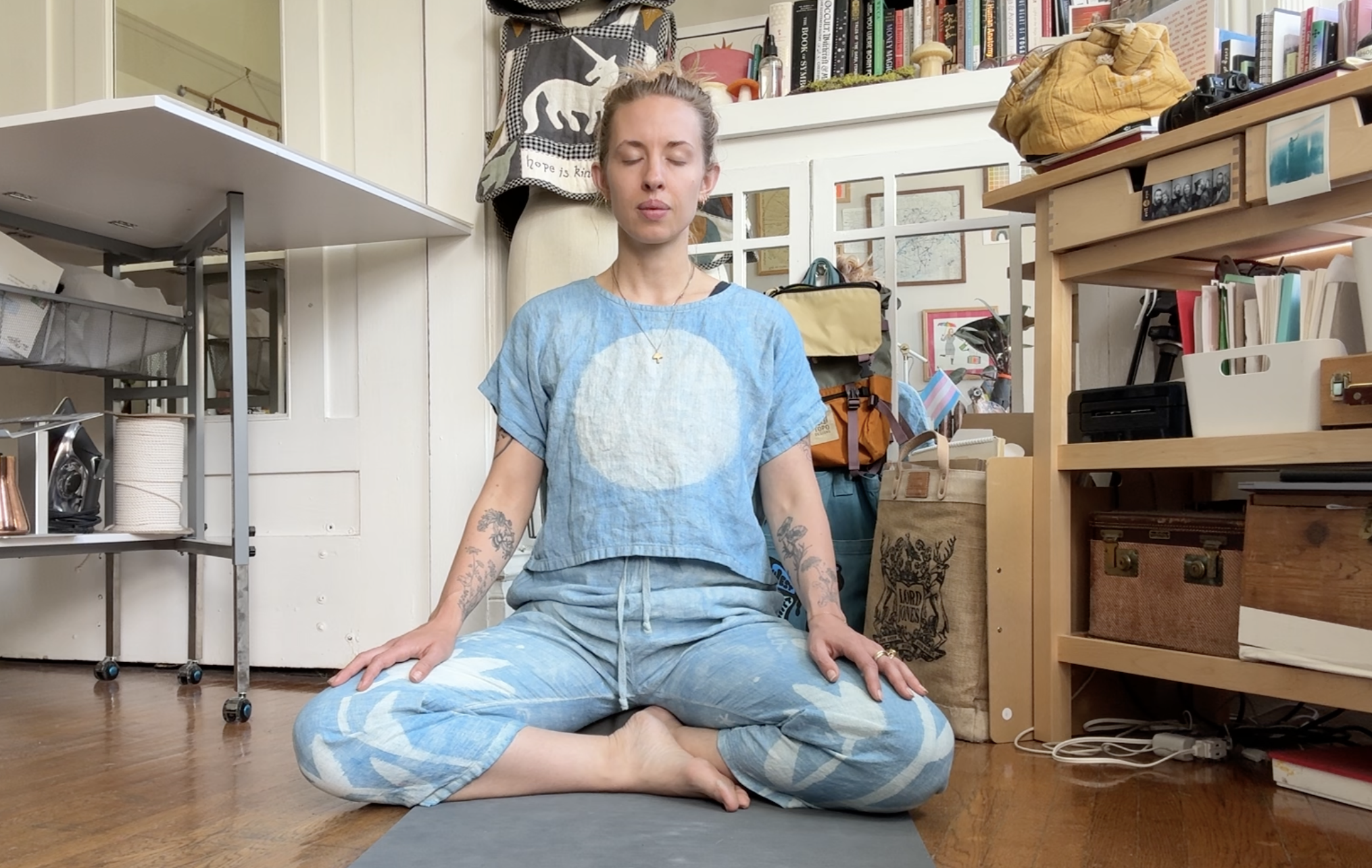 A woman with tattoos on her arms is sitting cross-legged on a yoga mat, in a room with shelves filled with books, bags, and various items. She is in a meditative pose with her eyes closed, wearing a blue tie-dye t-shirt and matching pants.