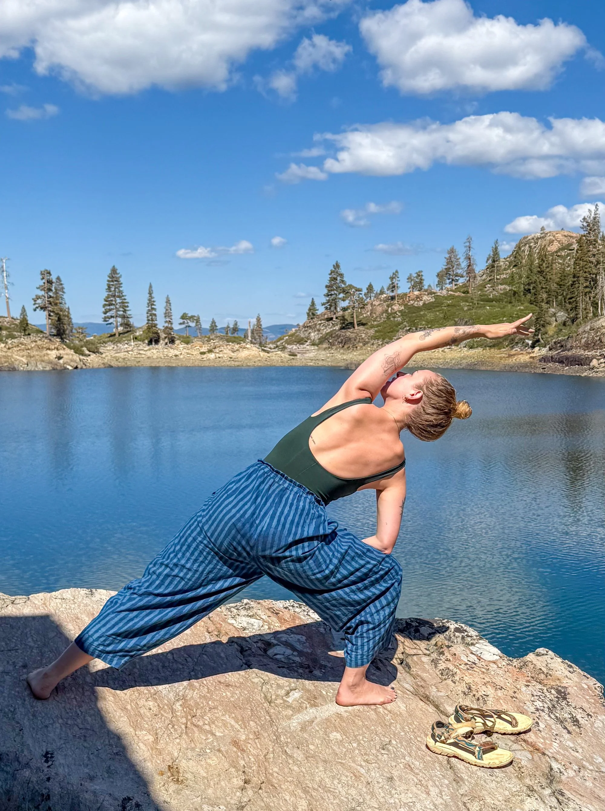 A woman in activewear performing yoga on a rock by a lake in a mountainous area.