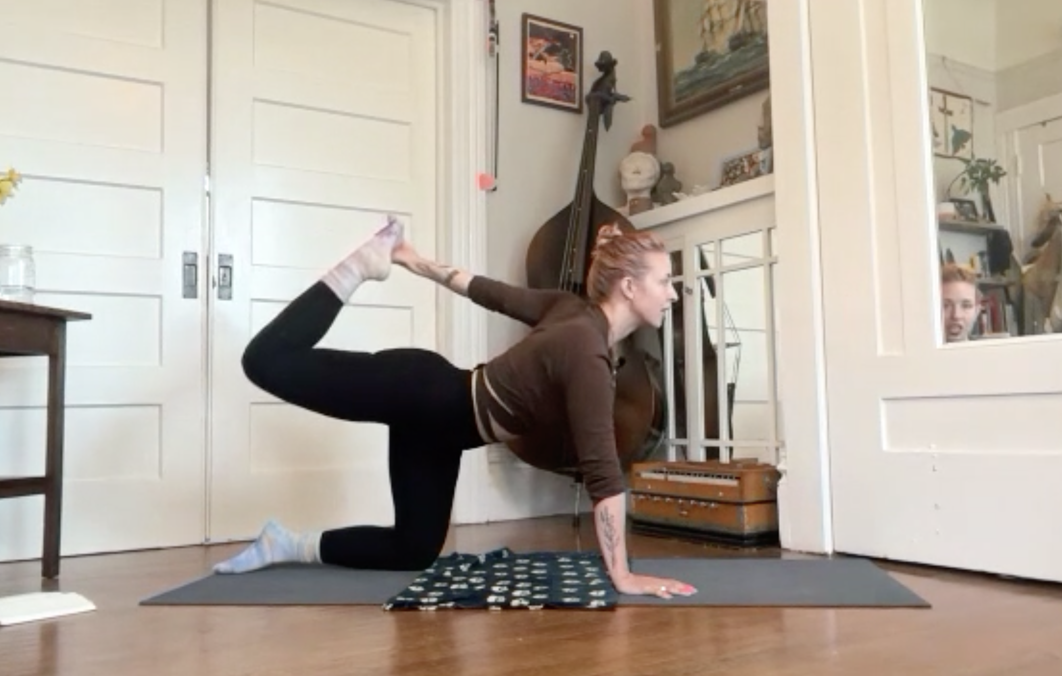 A woman with red hair practicing yoga indoors. She is balancing in a pose on her hands and knees, with one leg extended behind her and her head turned to the side. She is on a yoga mat in a room with white paneled closet doors, musical instruments, and framed artwork on the walls.