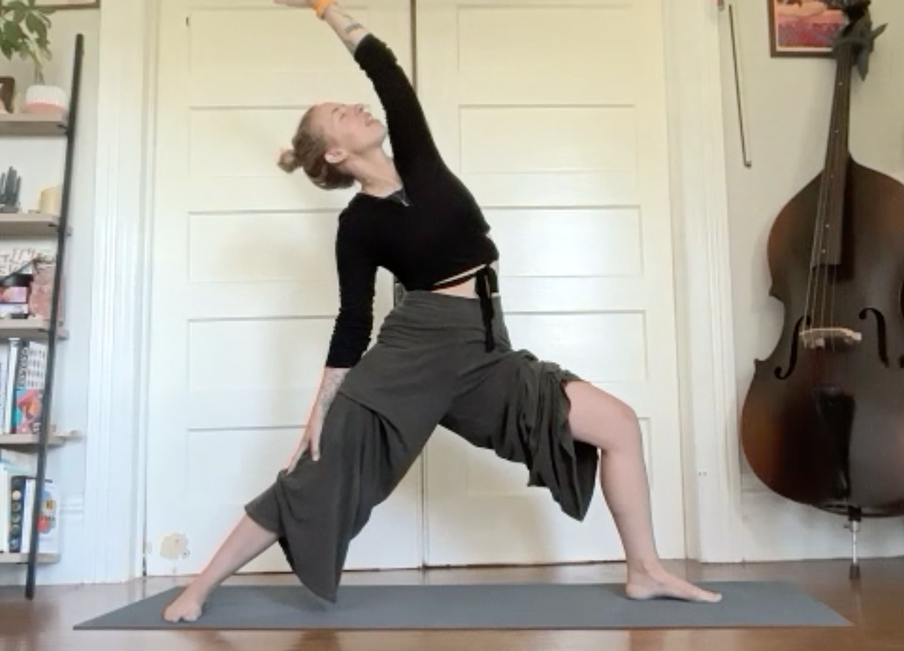 Woman practicing yoga in a lunge pose indoors, wearing a black crop top and loose gray pants, with a double bass leaning against the wall nearby.