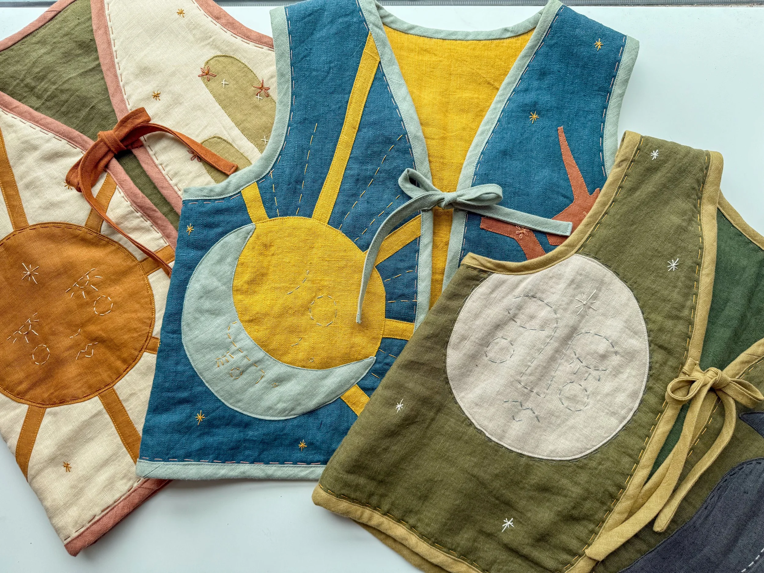 Colorful fabric bibs with embroidered moon and sun designs, featuring tie closures, laid flat on a white surface.