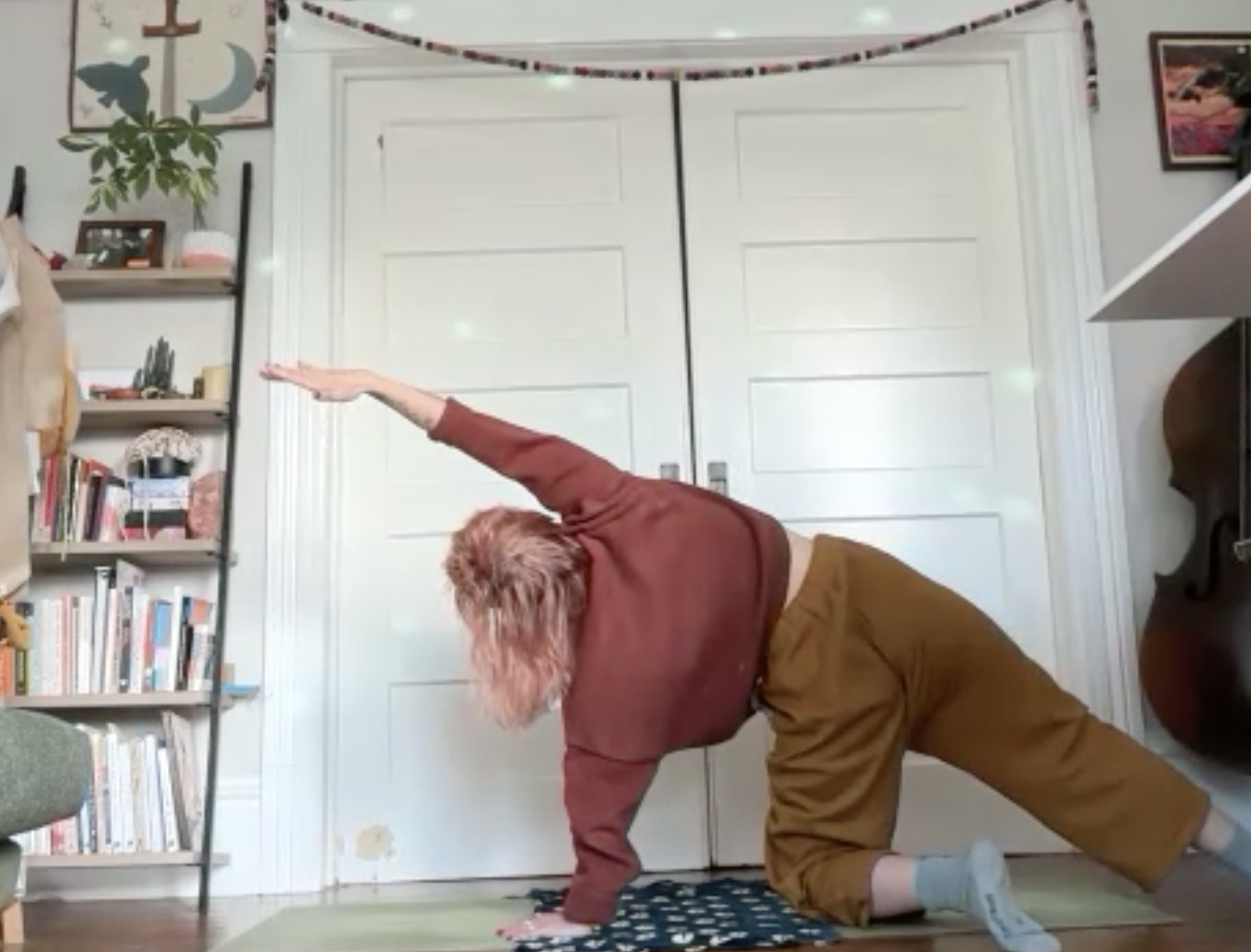 A person practicing yoga or stretching in a room with a bookshelf, plants, and framed pictures, performing a side bend with one arm extended overhead while on their knees.
