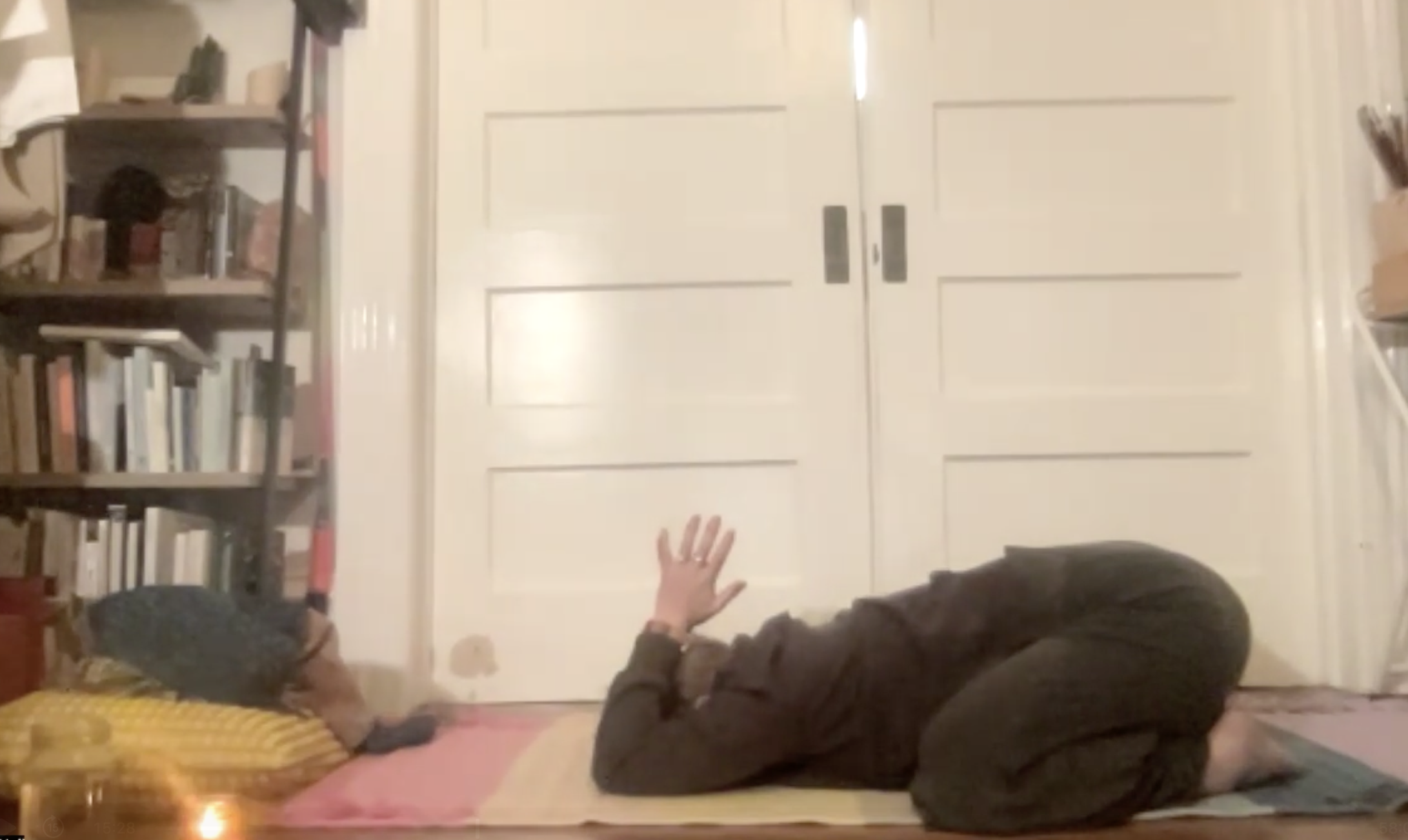 A person lying on the floor with arms outstretched toward another person who is seated nearby. The scene is in a room with a bookshelf on the left and closed double doors in the background.