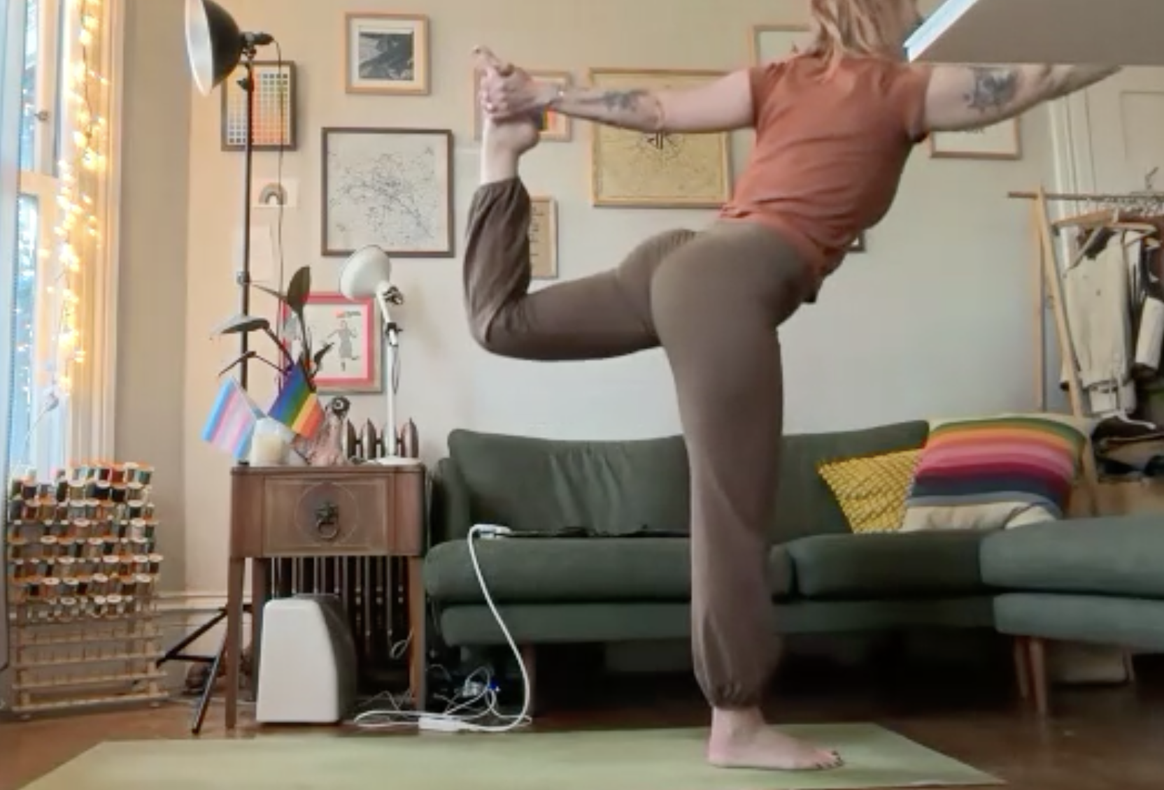A person in an indoor living room practicing yoga, standing on one leg with the other leg bent and foot held by one hand behind them. They are wearing a brown t-shirt and brown pants, with tattoos visible on their arms. The living room has a green couch, a rainbow-striped pillow, a framed picture gallery on the wall, and a window with string lights and pride flags.