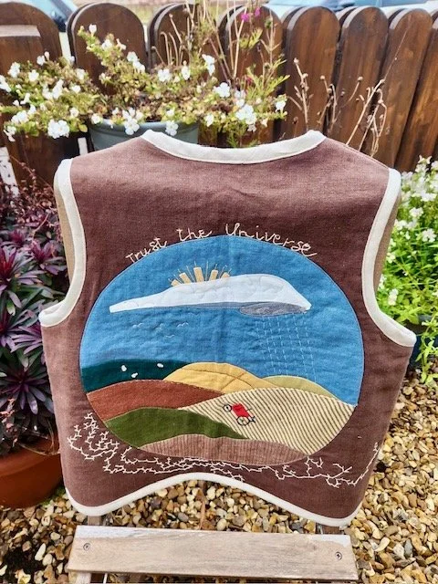 A quilted jacket with an embroidered design of a sunset and a landscape scene, with the words 'Trust the Universe' embroidered above, displayed outdoors against a wooden fence and surrounded by plants.