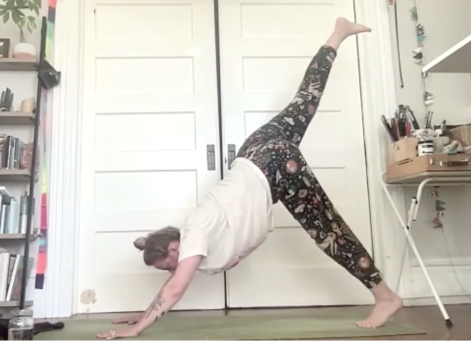 A woman practicing yoga indoors, performing a pose with one hand and one foot on the ground, and the other leg extended upward. She is on a green yoga mat in front of a white door, with bookshelves on the left and a storage shelf with art supplies on the right.