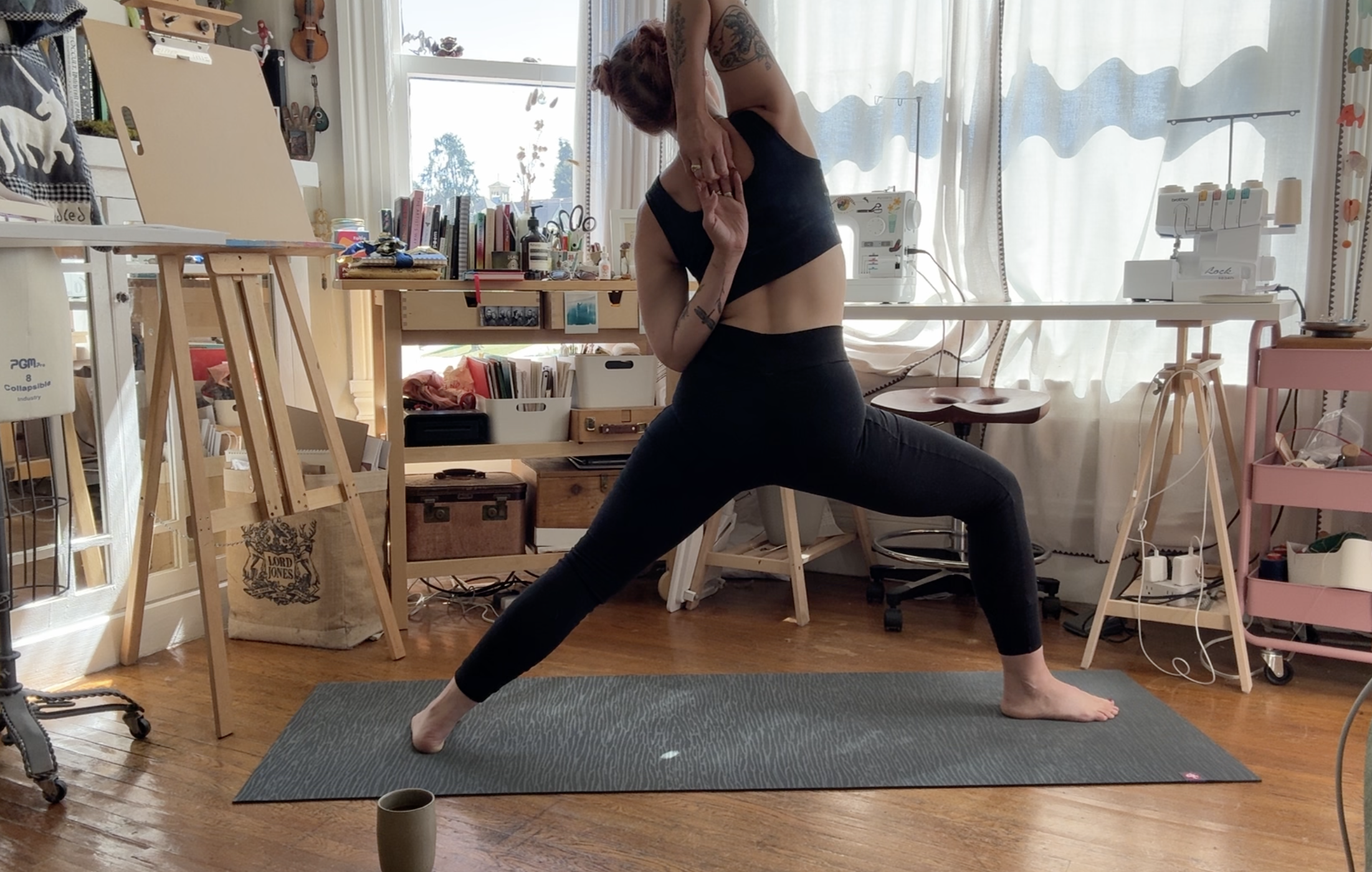 Woman practicing yoga indoors on a gray mat, in a lunge pose with arms raised, wearing black athletic wear, in a room with wooden floors and large windows.