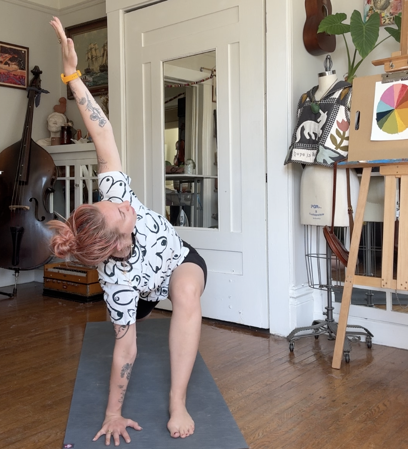 Person with pink hair in a house practicing yoga, in a pose balancing on one hand and foot with the other hand reaching upward, in a room with artwork, a sculpture, musical instrument, and art supplies.