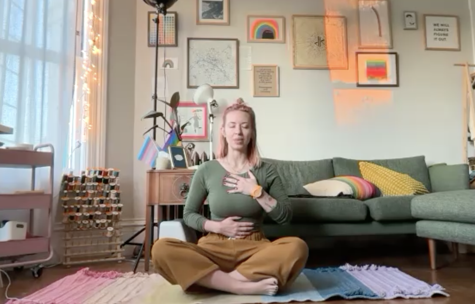 A woman with pink hair practicing meditation in a cozy living room. She is sitting cross-legged on a woven rug, with her hands on her chest and stomach closed eyes. The room is decorated with framed artwork on the wall, a gray couch with rainbow pillows, a small wooden cabinet, a floor lamp, and a collection of motivational messages and rainbow-themed decorations.